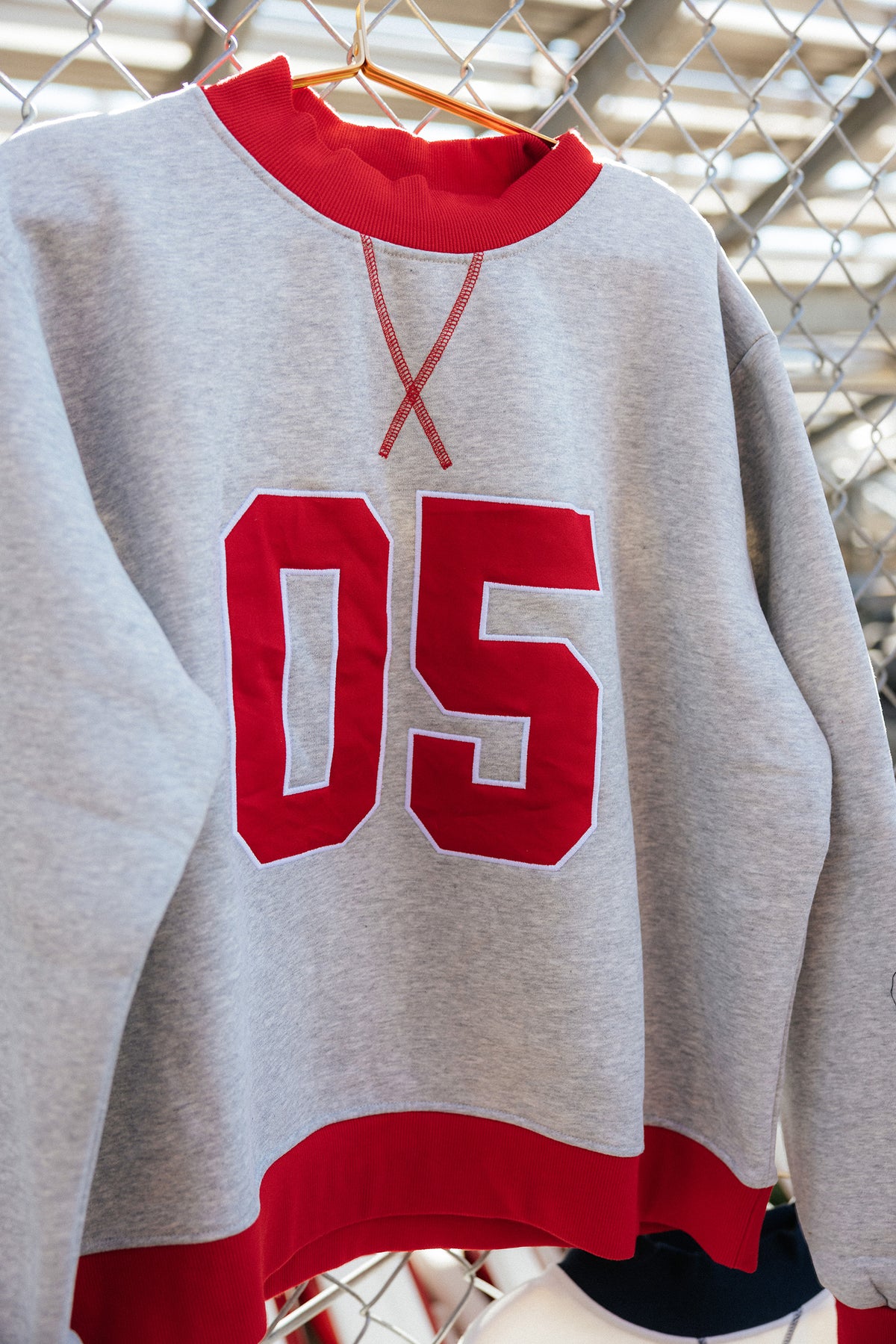 kids, A heather gray crewneck sweatshirt with red ribbed trim and large red '05' lettering on the front, hanging on a wooden hanger against a chain-link fence.