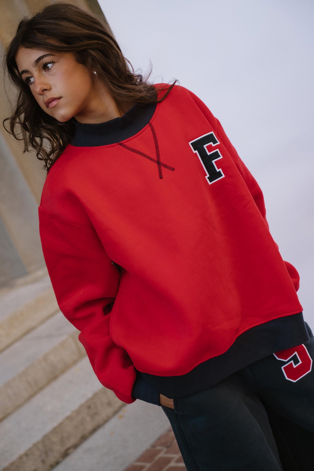 kids, A girl wearing a bright red crewneck sweatshirt with a navy ribbed collar, cuffs, and hem, featuring a navy collegiate letter 'F' on the left chest. She is also wearing navy sweats with a red and white '5' on the thigh.