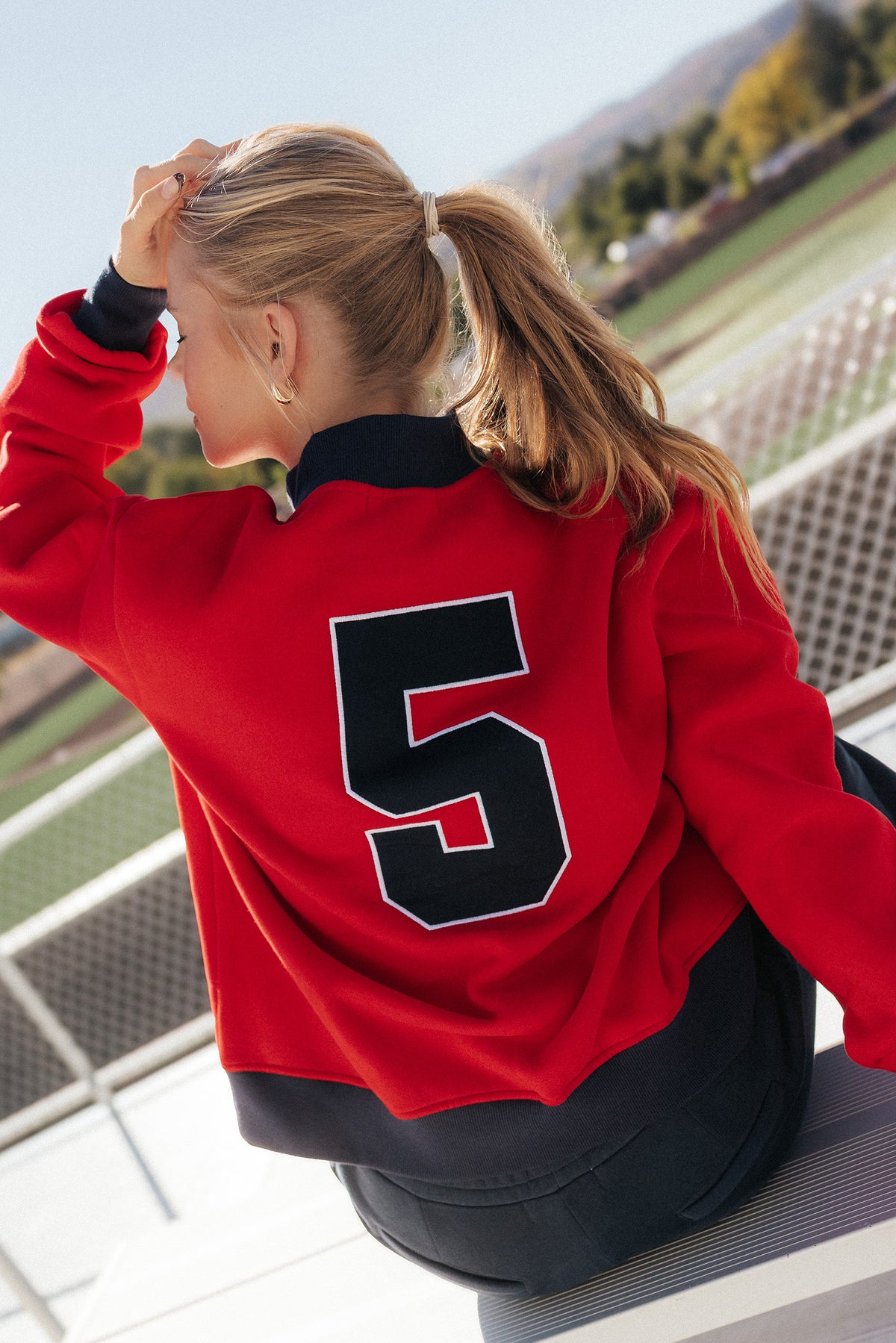 teen, A girl wearing a red and navy varsity-style sweatshirt with the number 5 on the back, photographed outdoors on a sunny day with a blurred field and mountains in the background.
