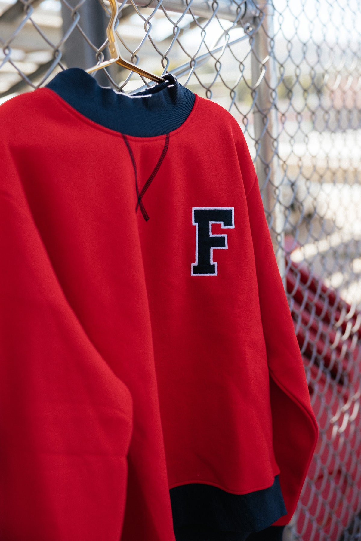teen, A red sweatshirt with a navy blue collar and hem, featuring a large navy blue letter 'F' with a white outline on the left chest, hanging on a wire fence.