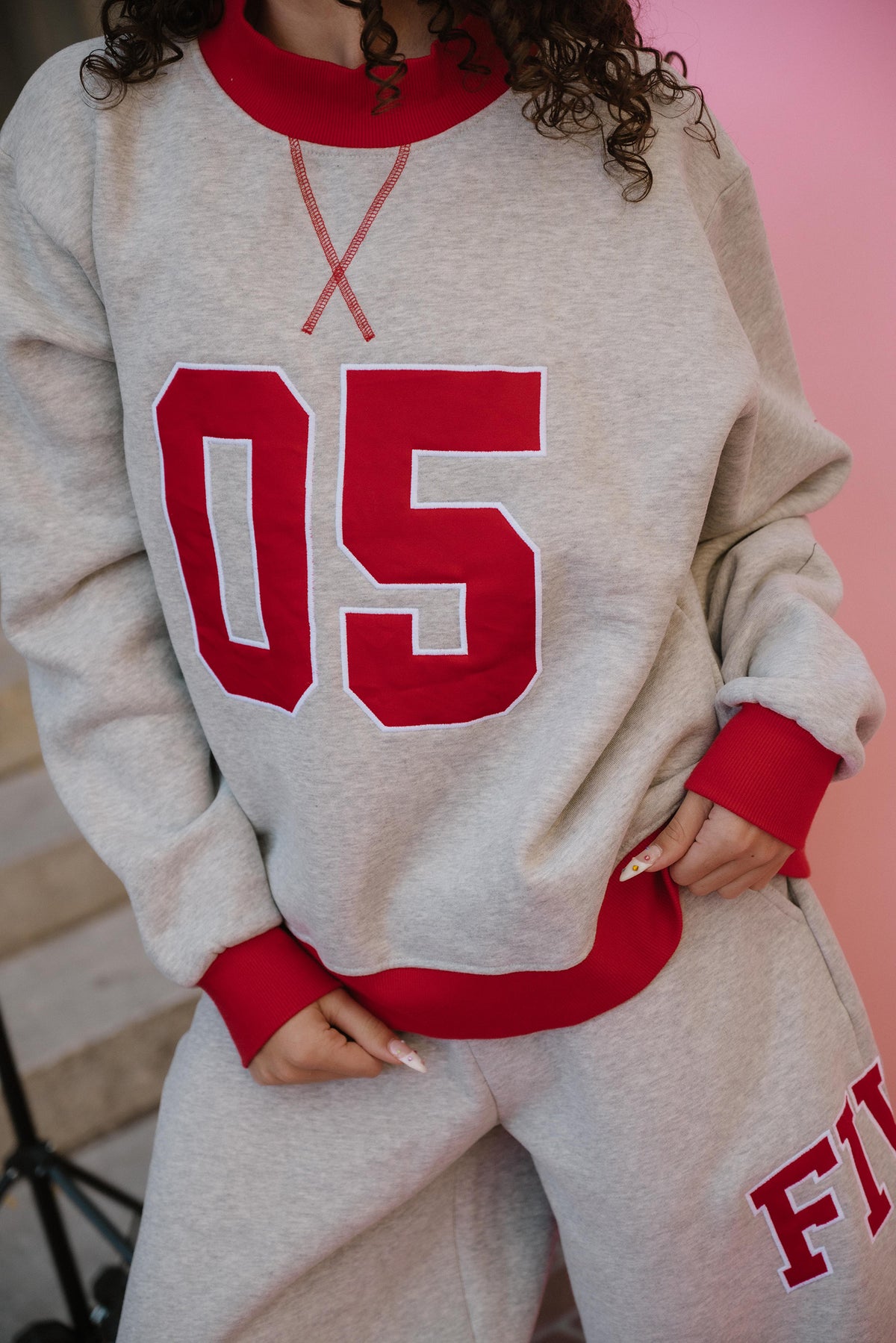 teen, A girl wearing a light heather gray sweatshirt and matching sweatpants set. The sweatshirt has a red crew neck, red ribbed cuffs and hem, and large red '05' lettering outlined in white on the front.