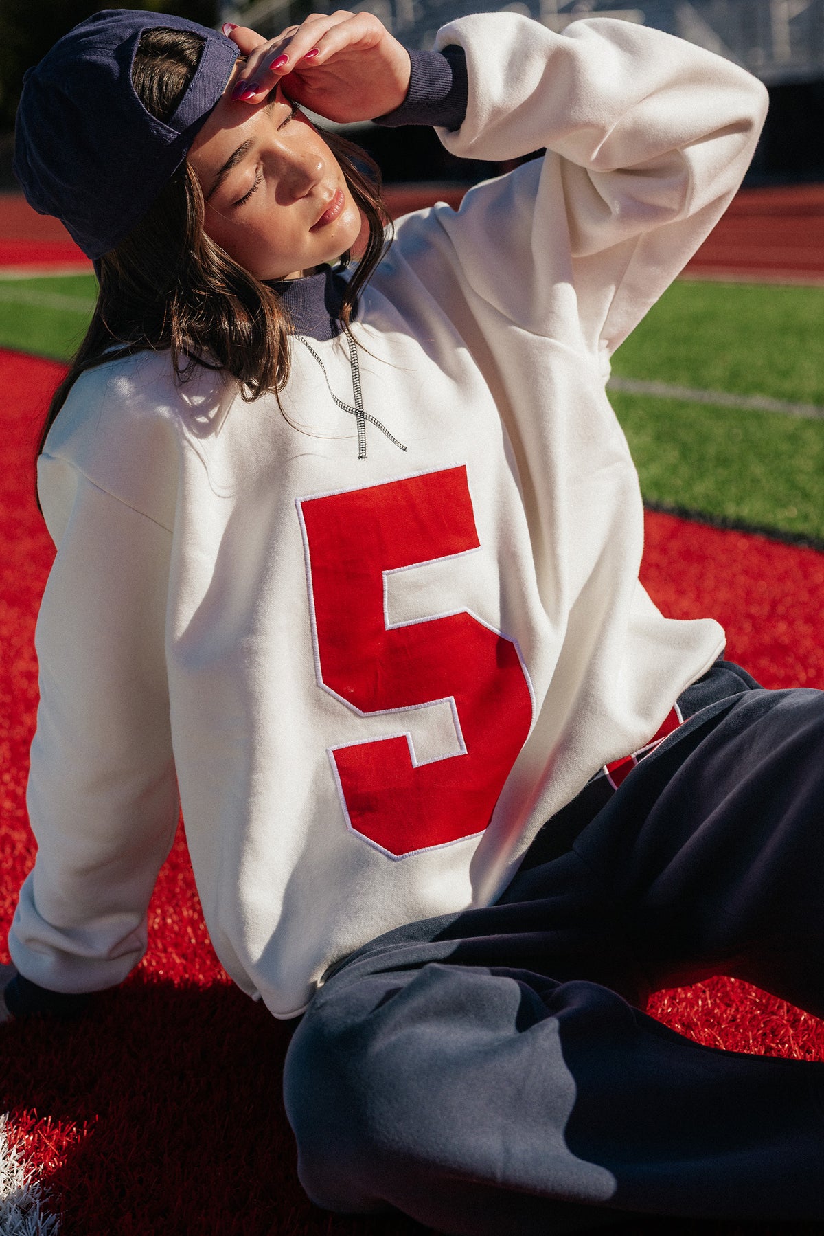 kids, sitting on a red turf field wearing an oversized cream sweatshirt with a large red number 5 on the front, navy sweatpants, and a navy baseball cap.
