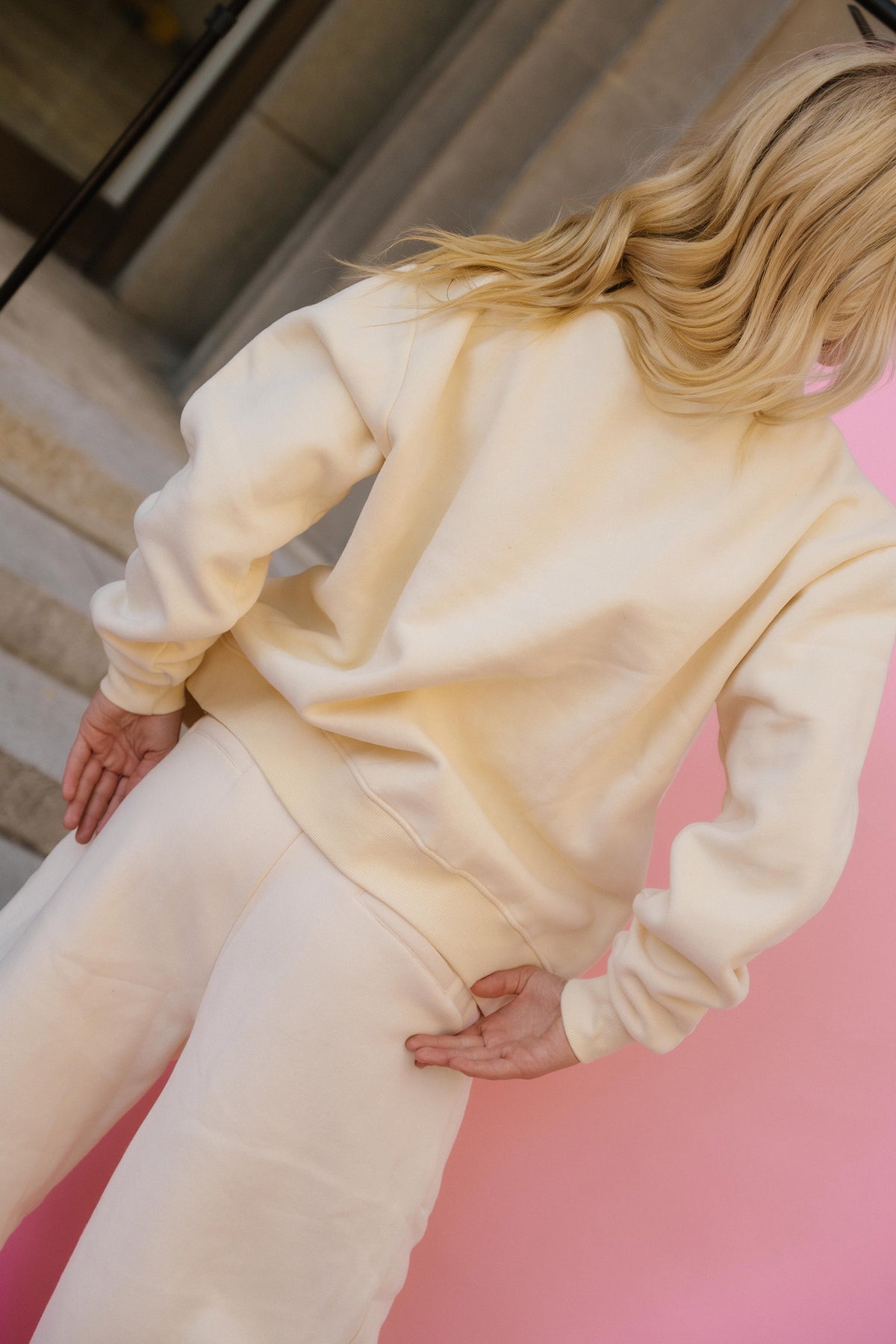 teen, wearing a butter-yellow colored sweatshirt and matching sweatpants, photographed from behind against a pink wall and stone staircase background.