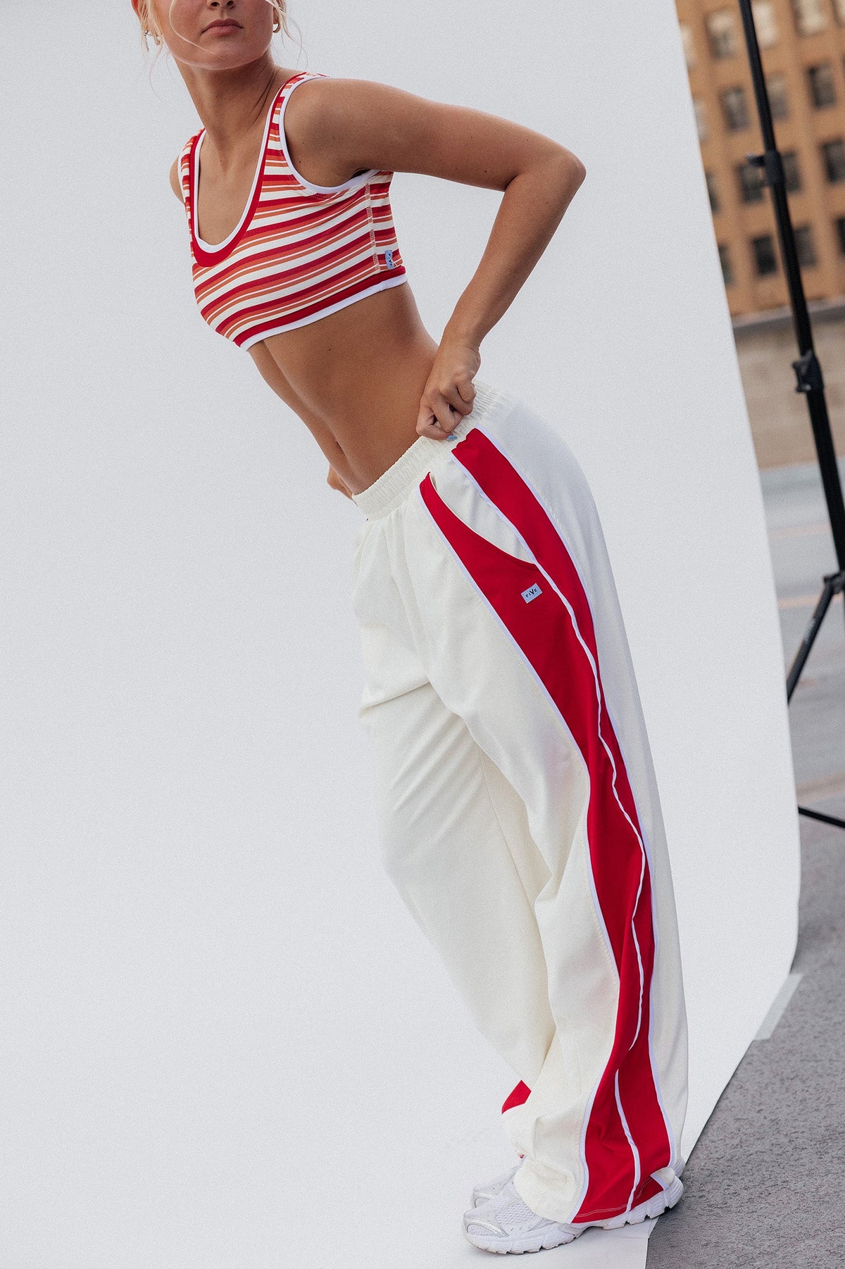 teen, girl posing in a red and white striped sports bra and white track pants with red side stripes, standing in front of a white studio backdrop with a city building visible in the background.