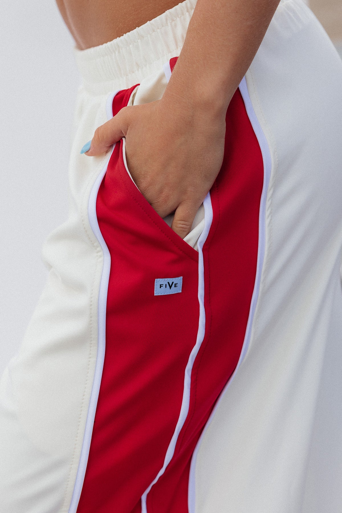 teen, Close-up of a girl wearing white athletic pants with a bold red side stripe and white piping, showing a hand in the side pocket. A small square logo patch reading 'five' is visible near the pocket.