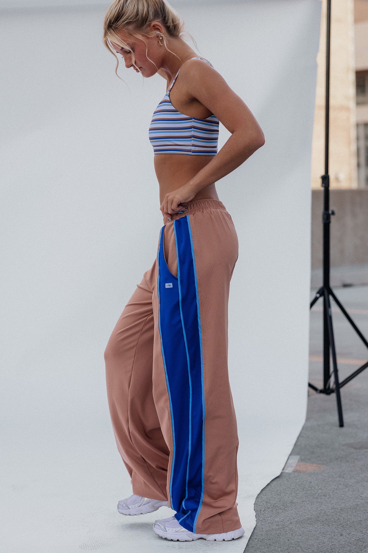 teen, girl stands in profile wearing wide-leg tan pants with a bold blue side stripe, a striped crop top, and white sneakers, posed in front of a white studio backdrop with a photography stand visible in an outdoor urban setting.