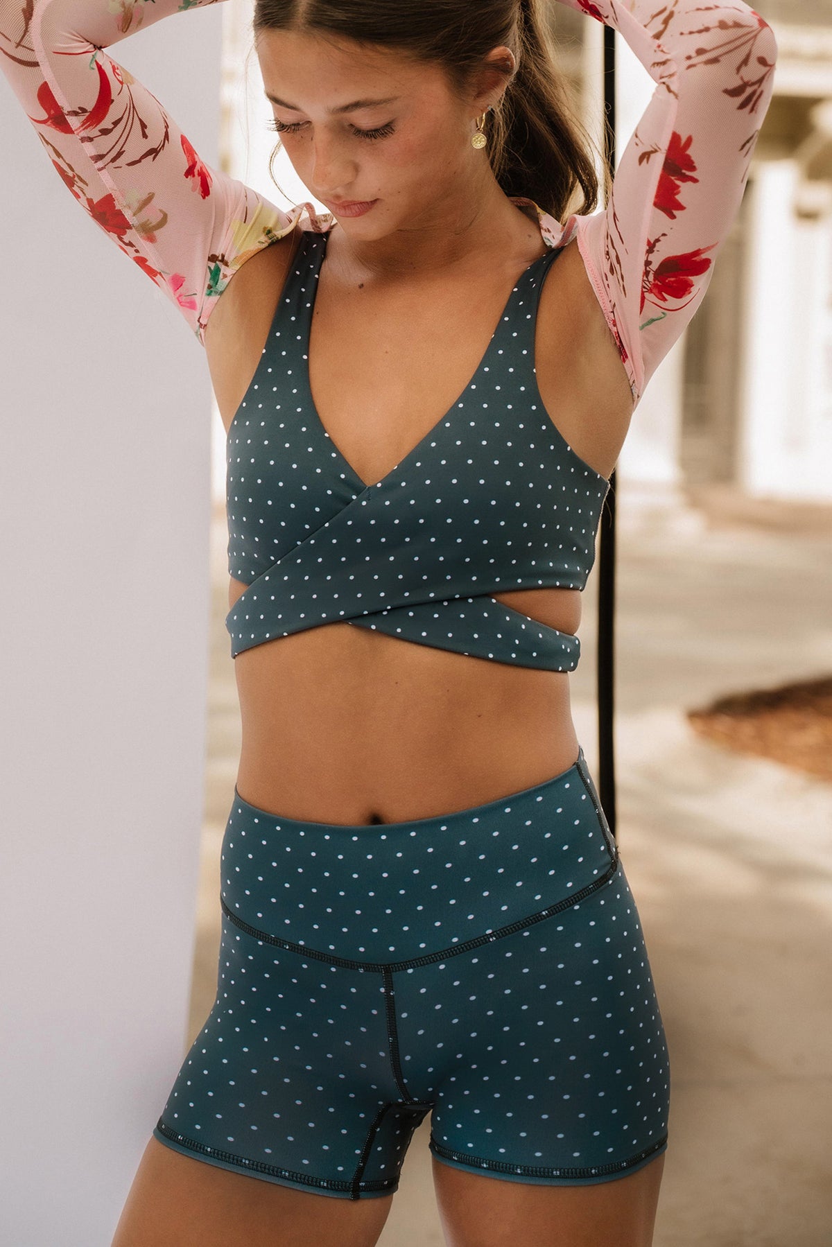 teen, modeling a matching dark blue sports bra and high-waisted shorts set with white polka dots, styled with a pink floral long-sleeve shirt in an outdoor setting.