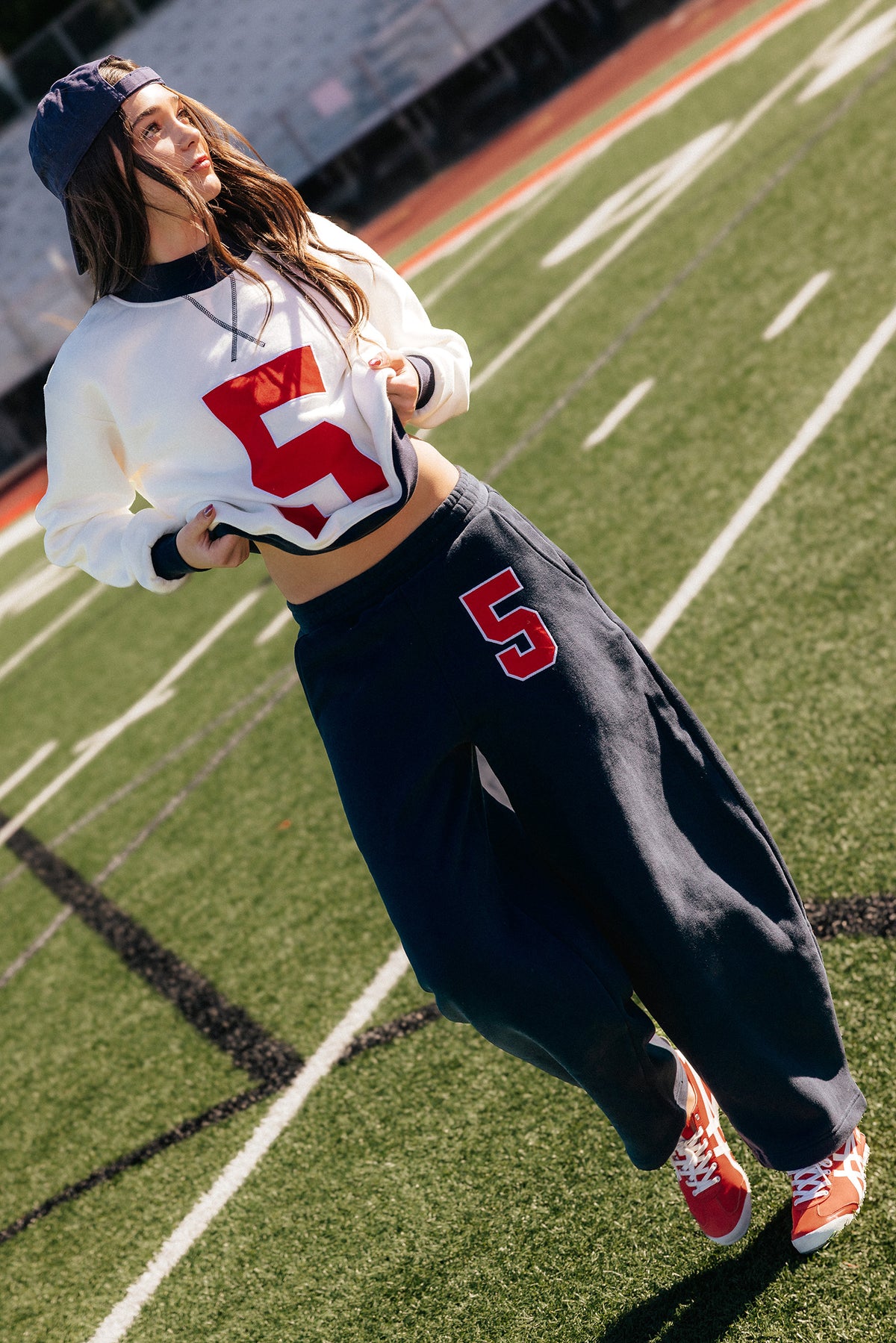 kids, girl poses on a green football field wearing a white cropped jersey with a red number 5, matching navy sweatpants, a navy cap, and red sneakers.