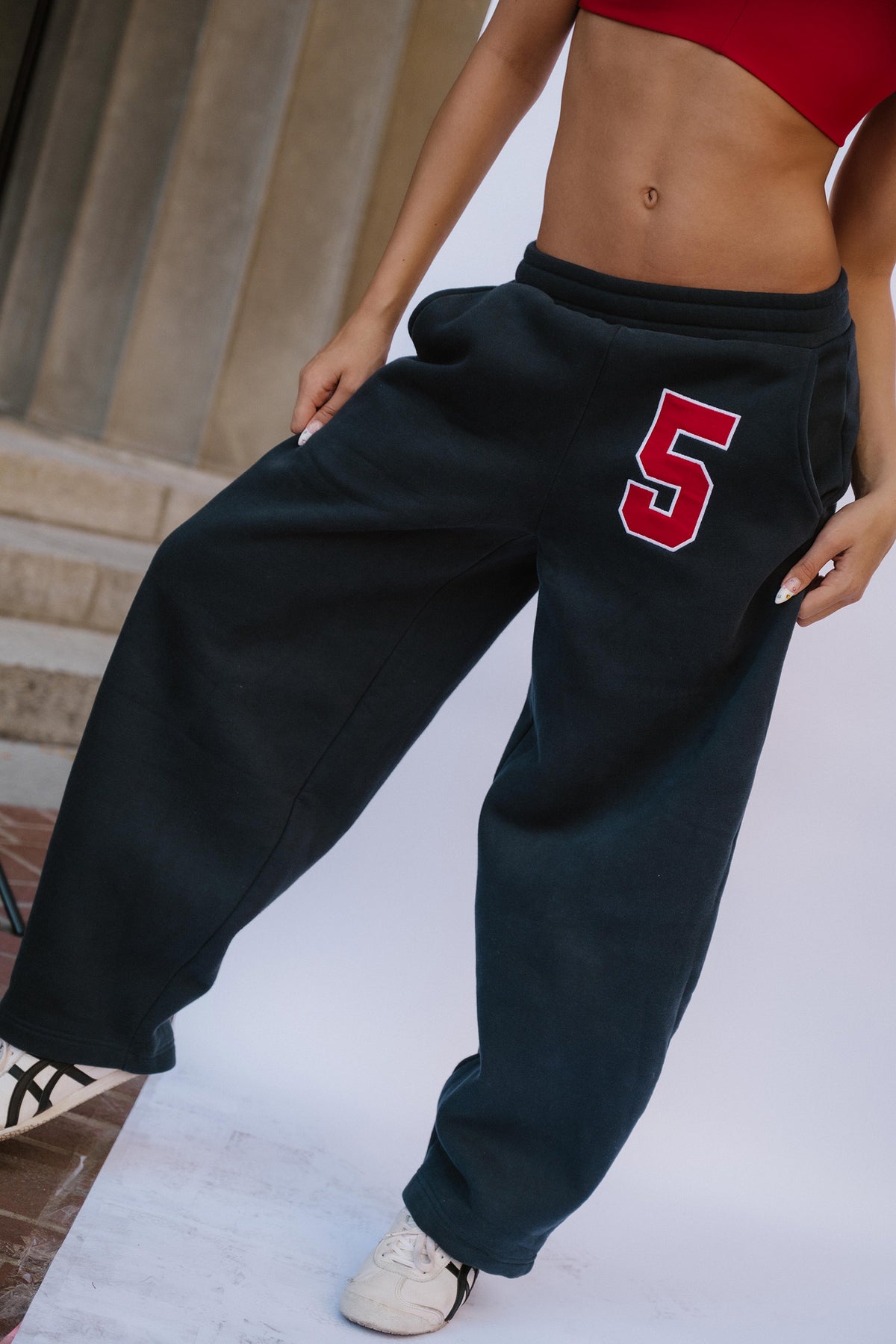 teen, wearing oversized dark navy sweatpants with a red number 5 on the left thigh, paired with a red crop top and white sneakers, standing in front of a white studio backdrop.