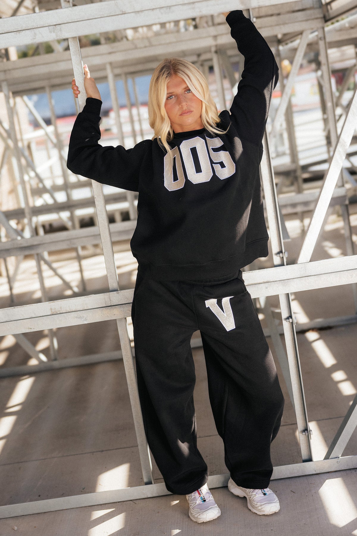teen, A girl posing in a black sweatshirt and sweatpants set with metallic silver varsity-style lettering, standing in front of a metal bleacher structure.