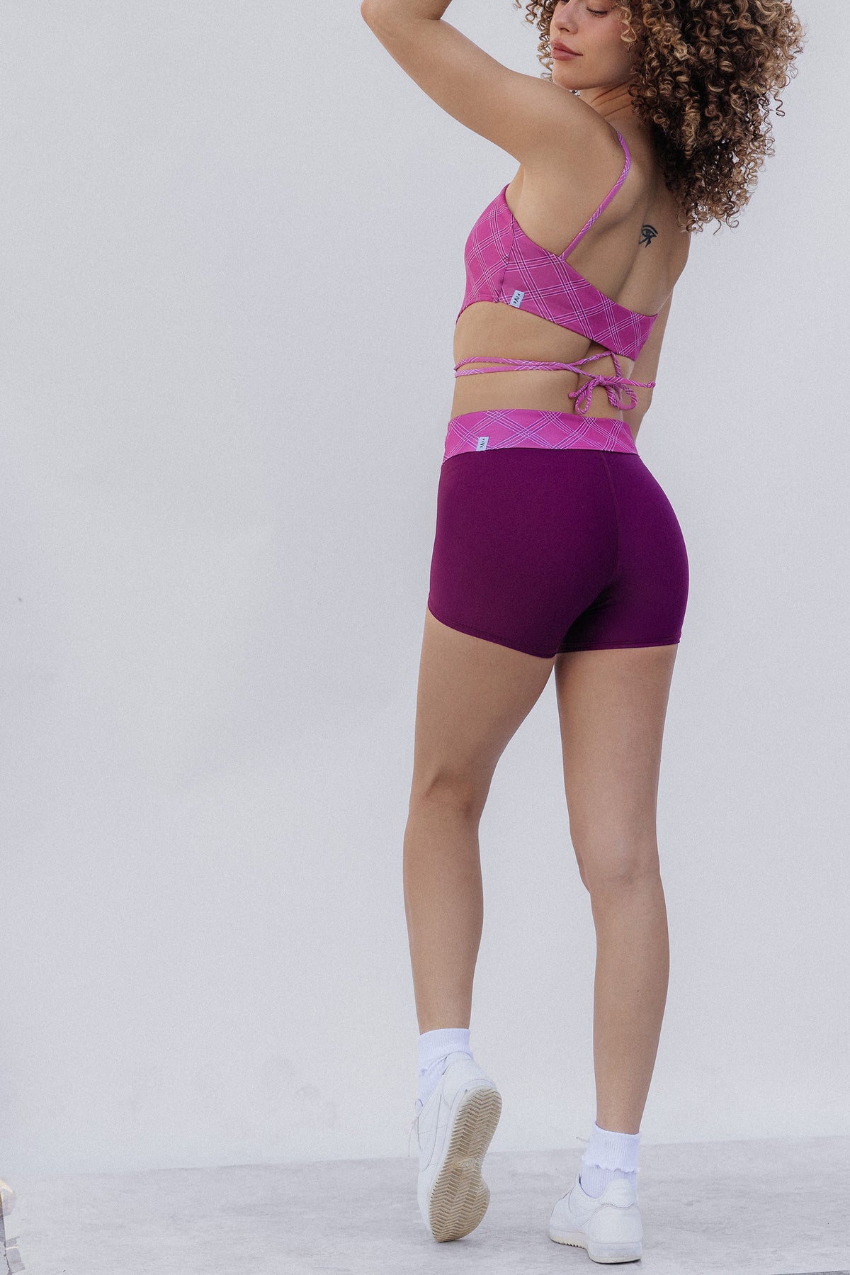 women, modeling a two-piece activewear set consisting of a pink and purple plaid sports bra and matching purple athletic shorts, shown from a rear three-quarter angle against a plain light gray studio background.