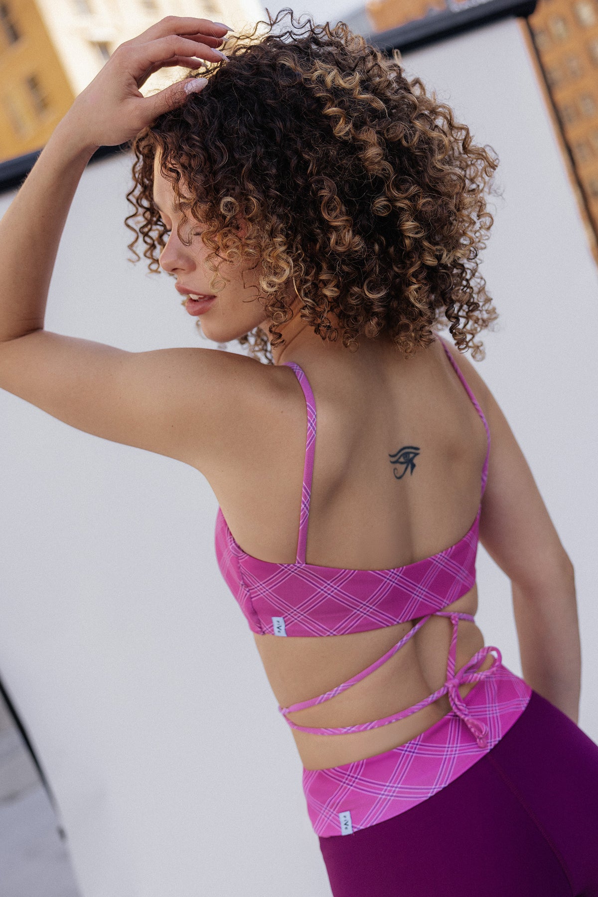 women, with curly brown and blonde hair is shown from the back wearing a pink plaid sports bra and matching purple leggings. She is posing on a white wall and buildings in the background.