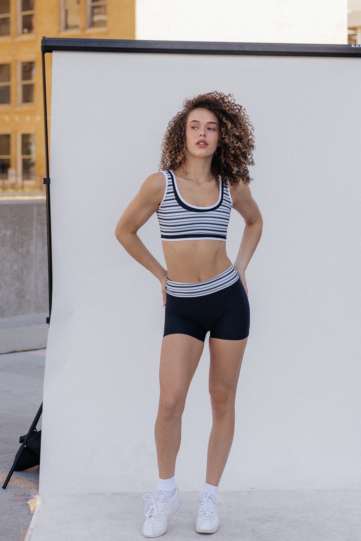 women, A woman stands in front of a white studio backdrop wearing a black and white horizontally striped sports bra and matching high-waisted athletic shorts, paired with white socks and sneakers.