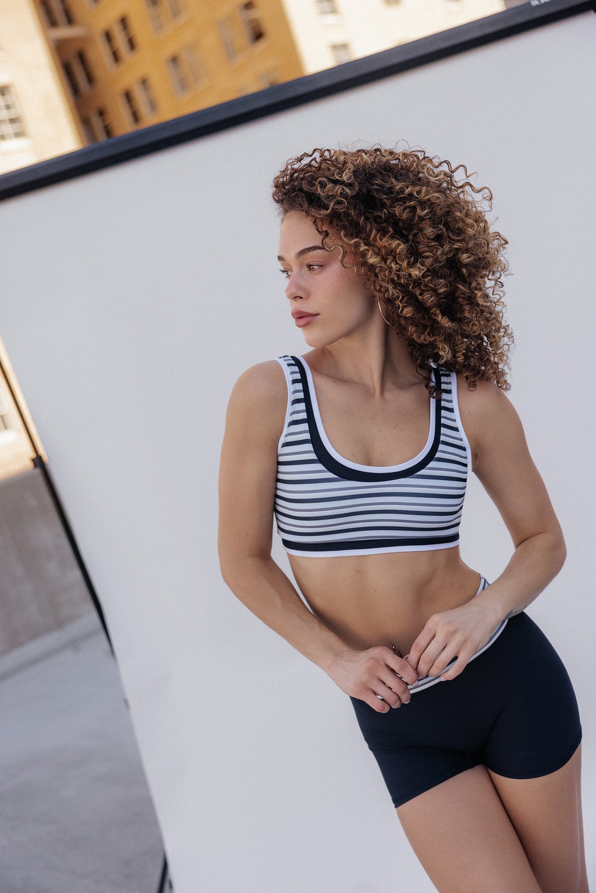 women, A woman with curly brown hair wearing a black and white striped sports bra and black athletic shorts, posing outdoors against a white wall.