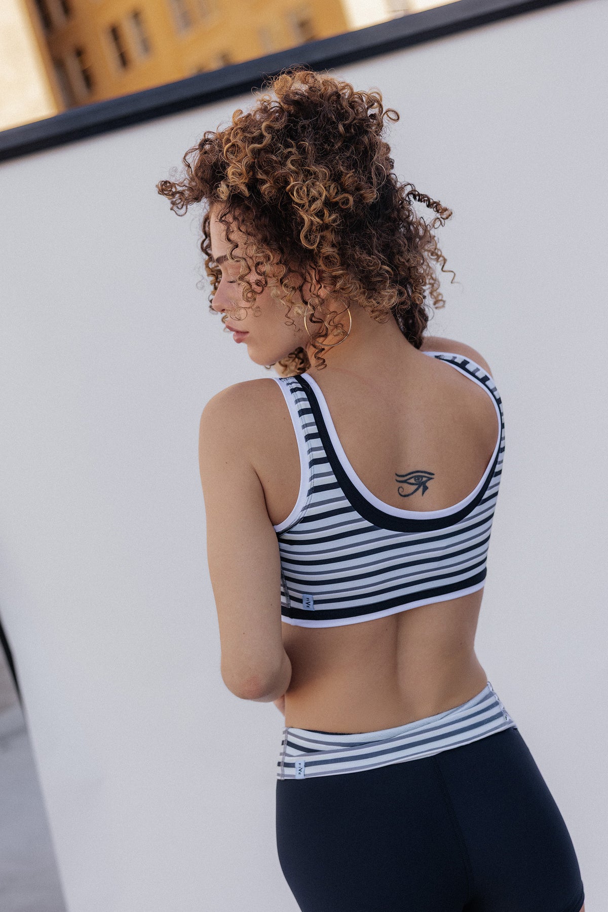 women, A woman with curly brown hair is shown from the back wearing a black and white striped athletic sports bra and dark leggings against a white wall.