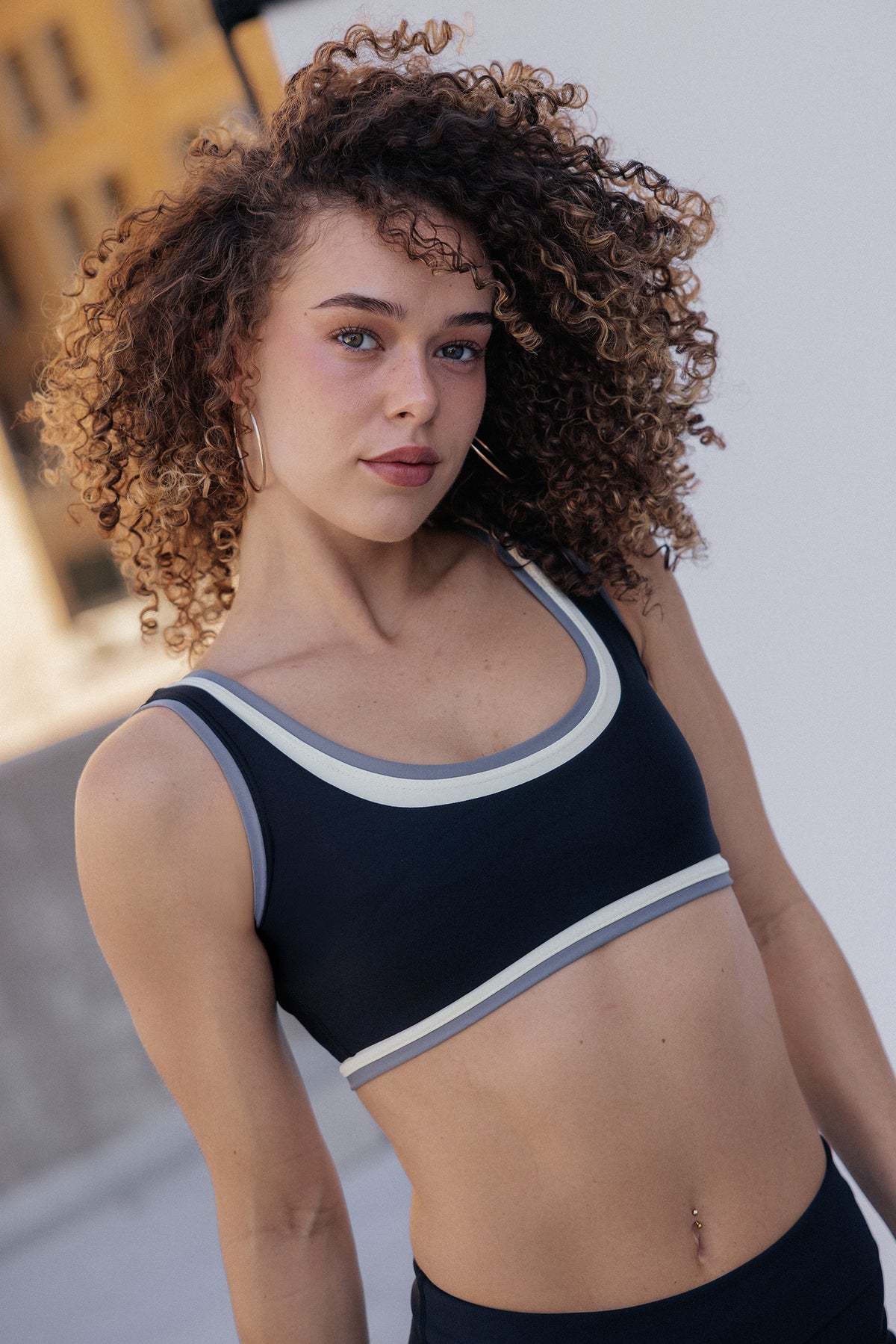 women, A woman with voluminous curly brown hair wearing a black sports bra with white and gray trim, photographed outdoors against a light gray wall.