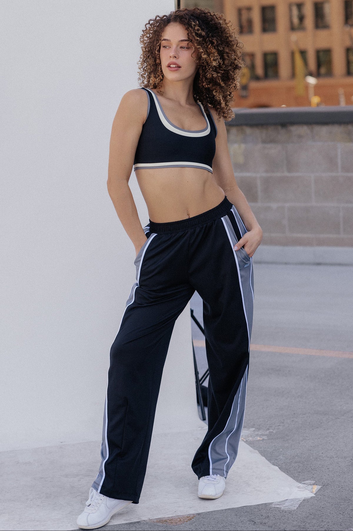 A woman with voluminous curly brown hair wearing a black sports bra with white and gray trim, photographed outdoors against a light gray wall.