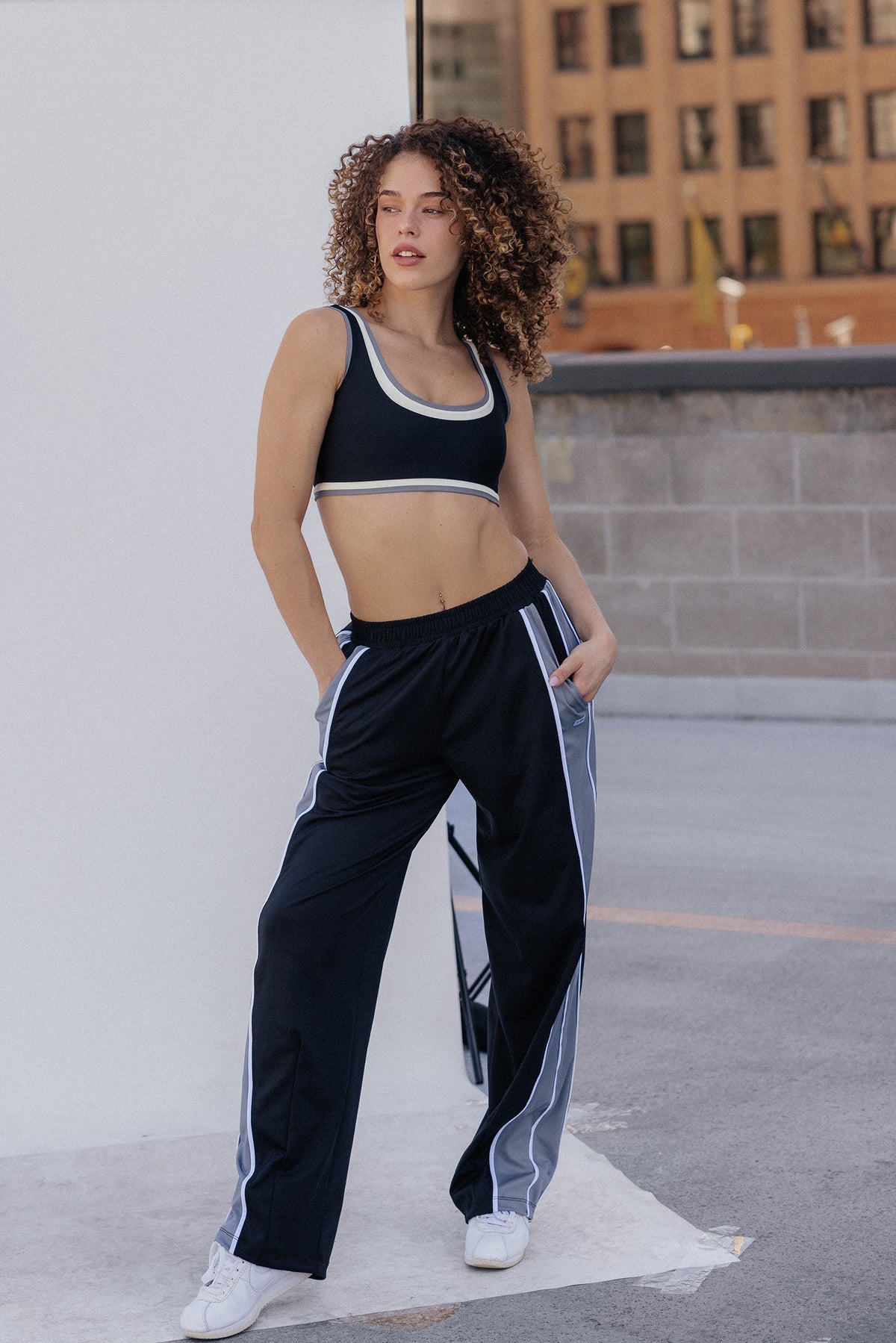 women, A woman with voluminous curly brown hair wearing a black sports bra with white and gray trim, photographed outdoors against a light gray wall.