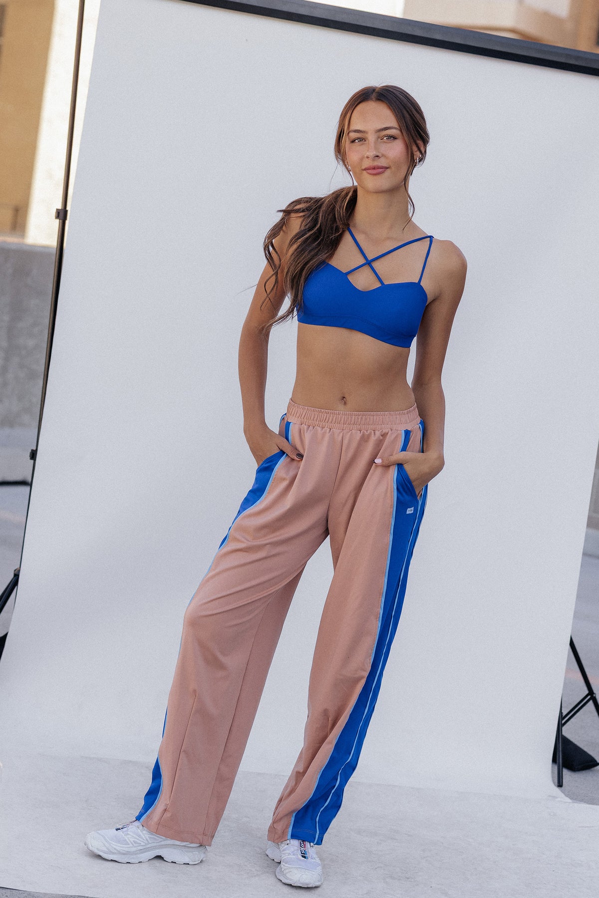 women, A woman modeling a two-piece activewear set consisting of a royal blue strappy sports bra and nude color wide-leg track pants with blue side stripes, standing in front of a white studio backdrop.