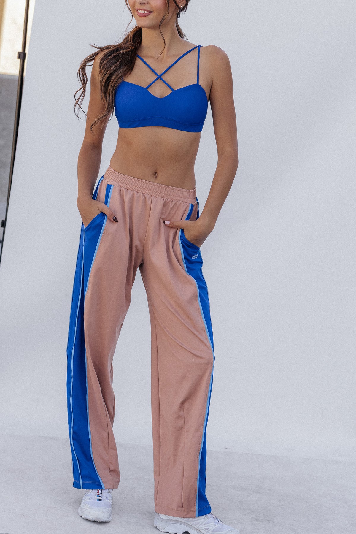 women, A woman modeling a two-piece activewear set consisting of a royal blue strappy sports bra and nude color wide-leg track pants with blue side stripes, standing in front of a white studio backdrop.
