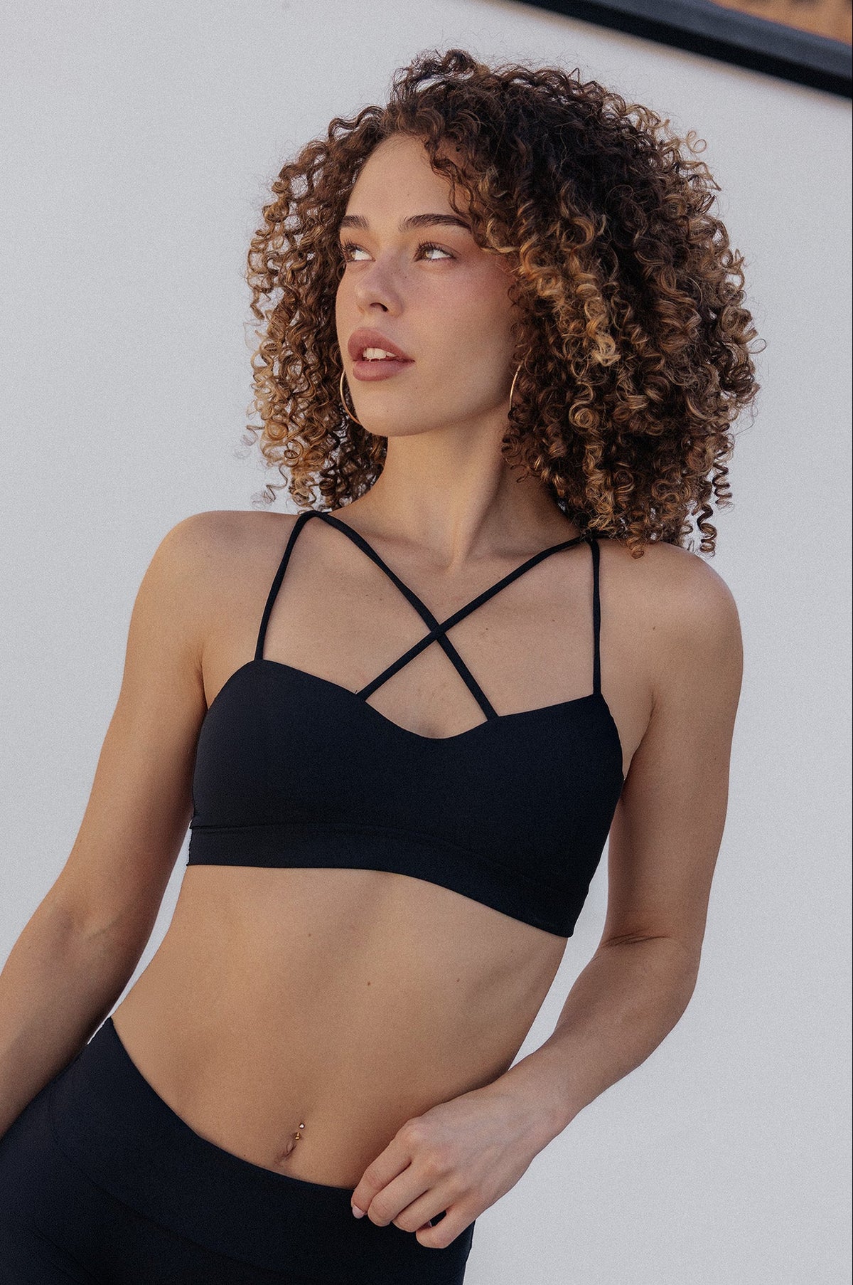 A woman with curly brown hair wearing a black strappy sports bra and matching black athletic shorts, posing against a white wall.