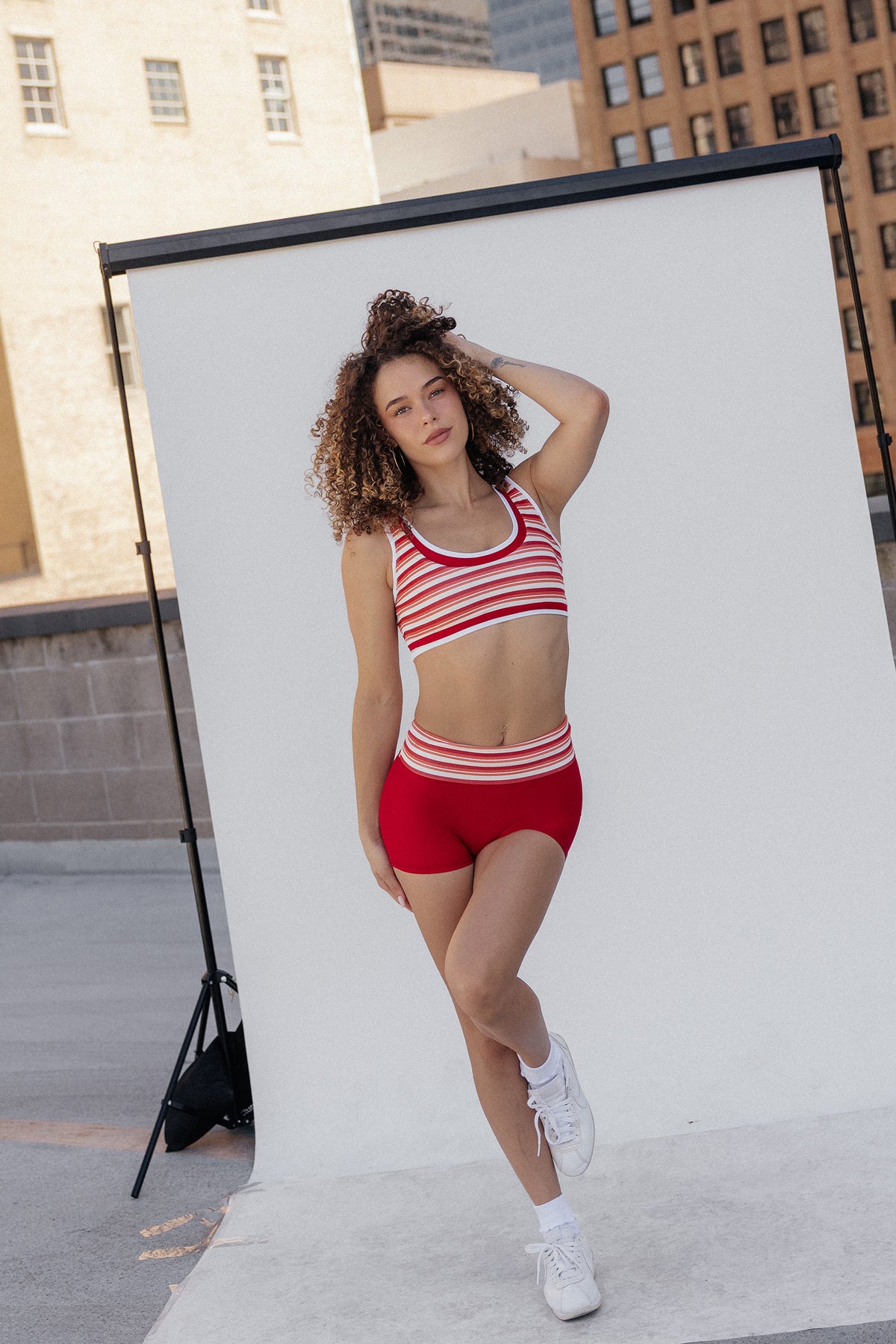 women, A woman with curly brown and blonde hair is shown from behind wearing a red and white striped sports bra and matching red bottoms against a light gray background.