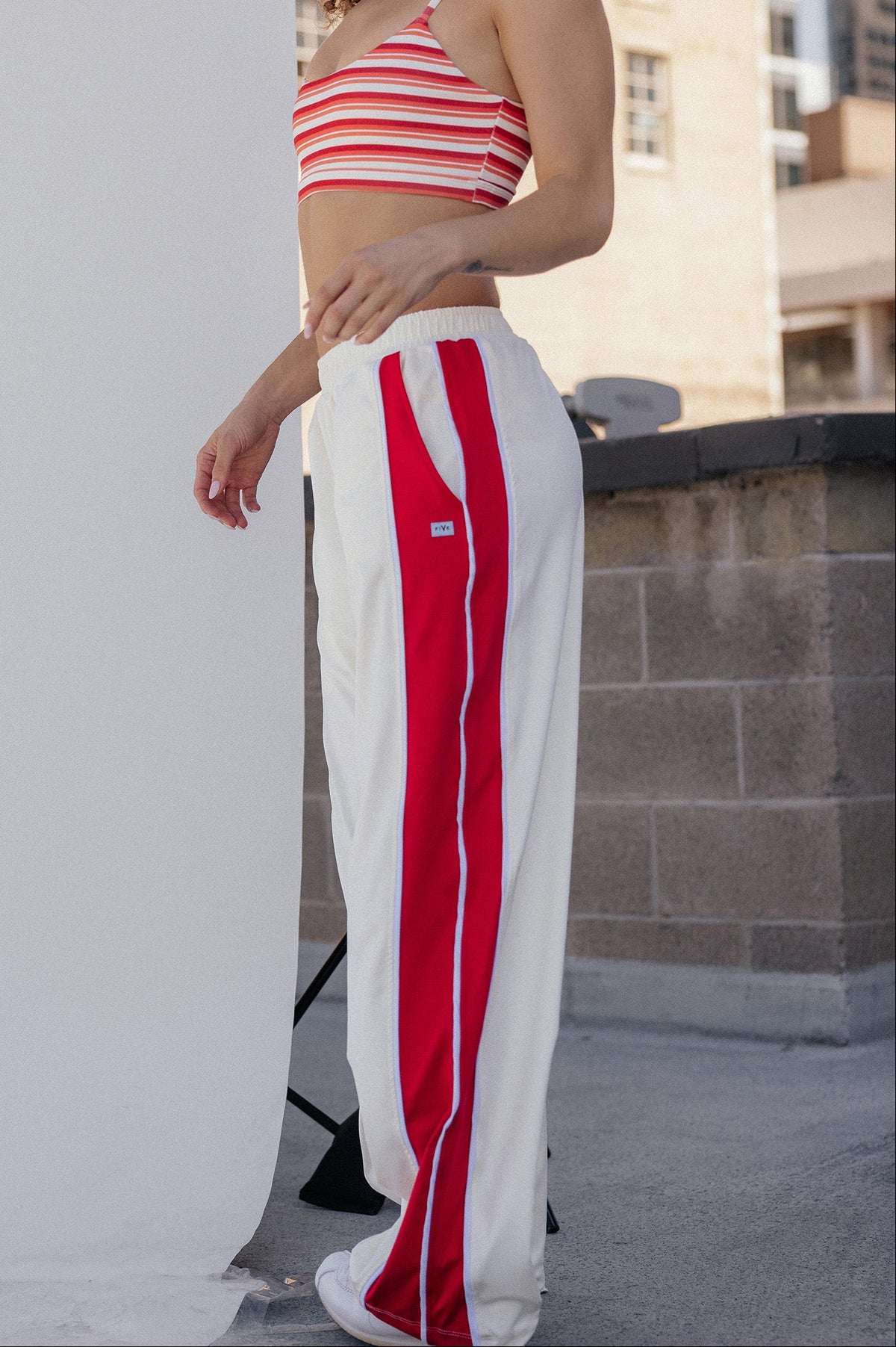 A model wearing a red and white striped crop top and matching red and white wide-leg track pants, standing outdoors next to a white studio backdrop.