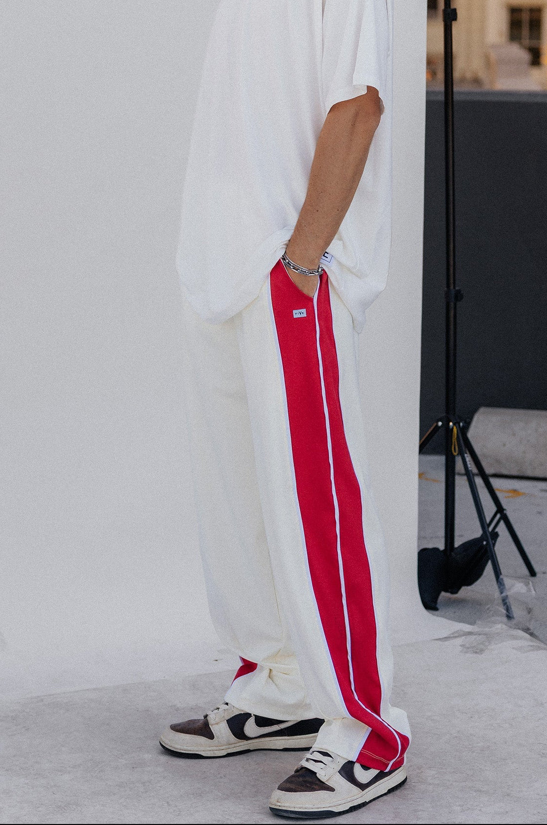 men, A man stands in a studio setting wearing a loose white t-shirt, white sweatpants with a bold red side stripe, and white sneakers with red and black accents. A large white backdrop screen is behind him, with a city building visible in the background.