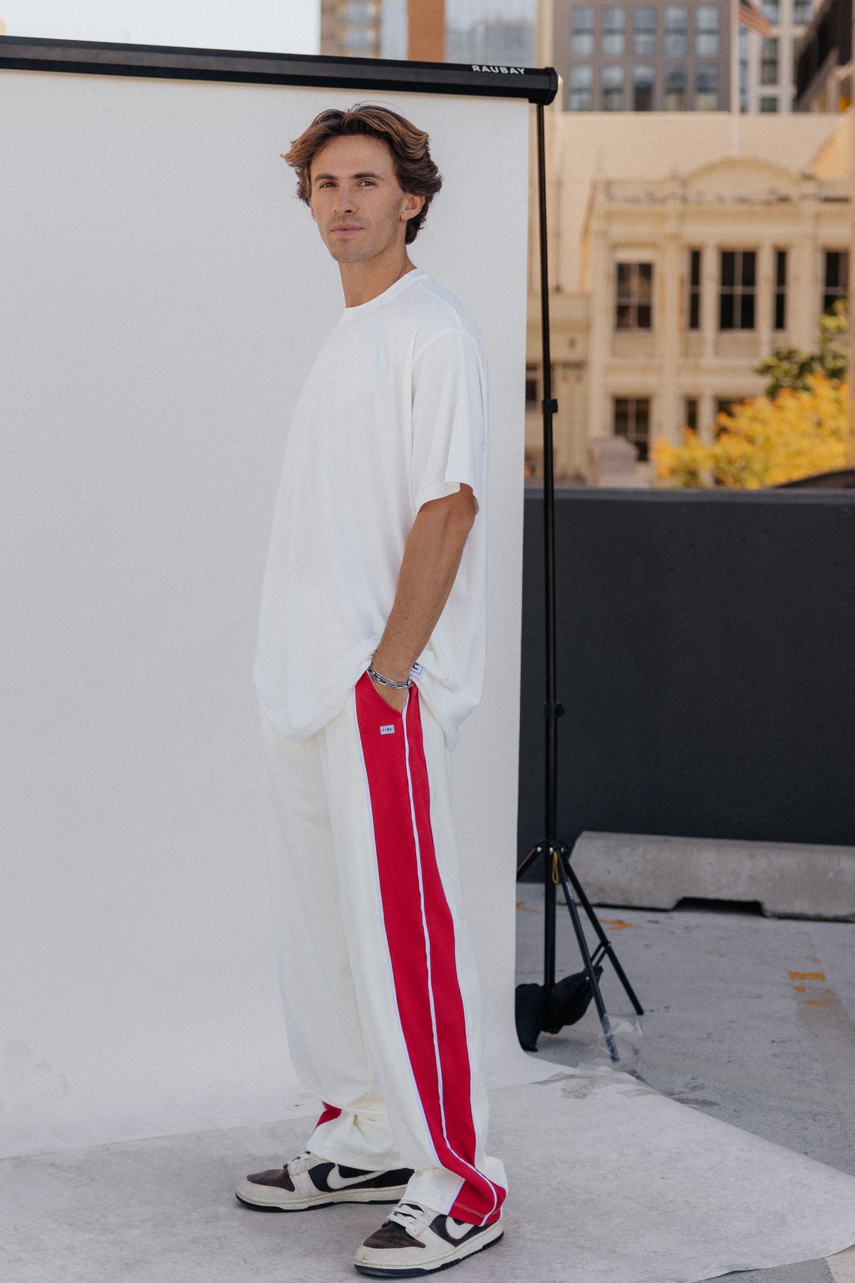 men, A man stands in a studio setting wearing a loose white t-shirt, white sweatpants with a bold red side stripe, and white sneakers with red and black accents. A large white backdrop screen is behind him, with a city building visible in the background.