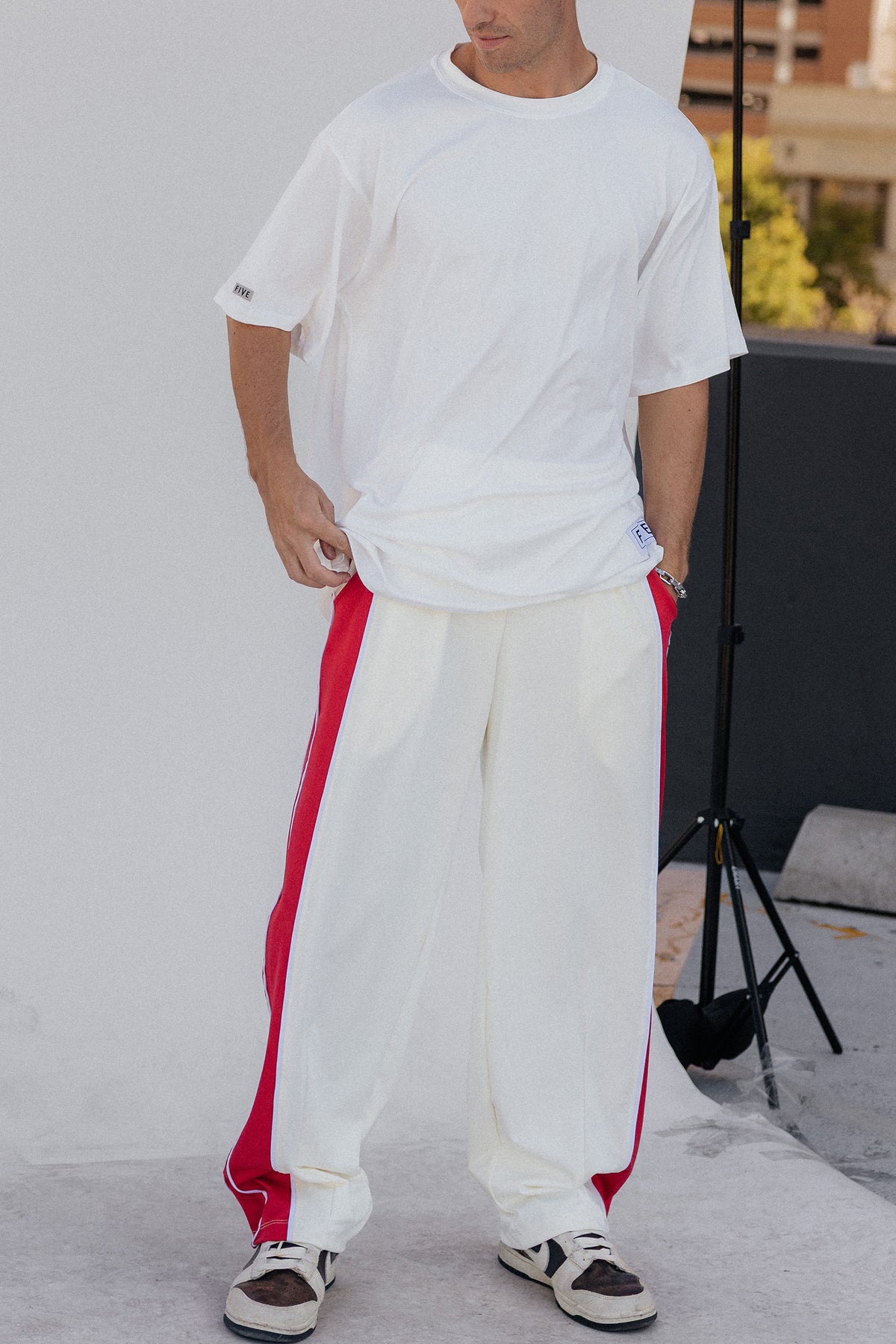 men, A man wearing a white oversized t-shirt and white sweatpants with red side stripes, paired with white and black sneakers, standing against a light gray wall.

