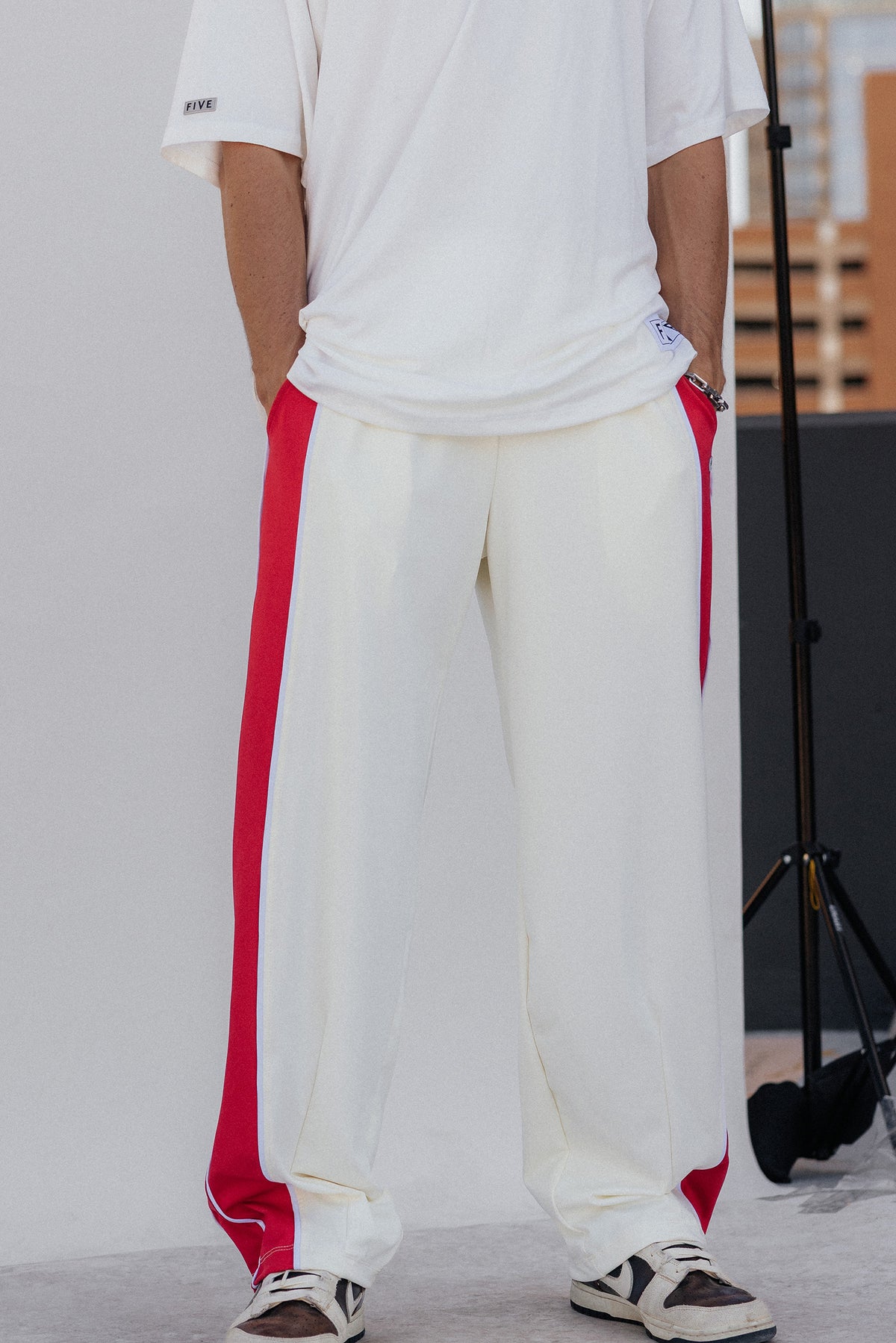 men, A person wearing loose white side-slit track pants with bold red side panels, paired with a white T-shirt and white sneakers, photographed indoors against a light gray backdrop.