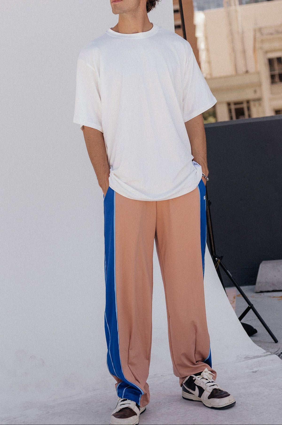 men, A man modeling a white oversized t-shirt and nude-colored track pants with blue side stripes, standing in front of a white studio backdrop with a city skyline in the background.