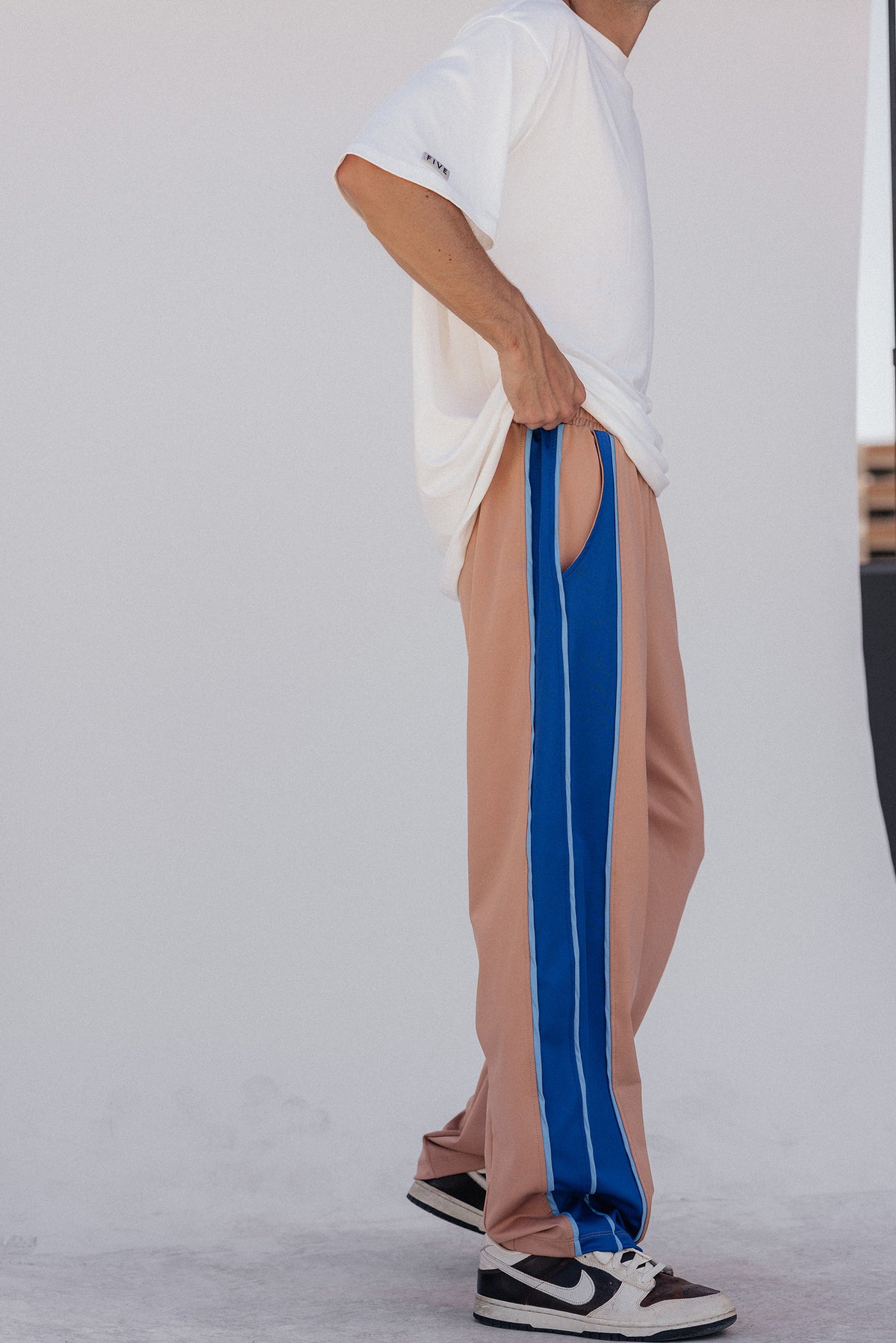 men, A person wearing loose nude track pants with bold blue side stripes, paired with a white oversized T-shirt and black-and-white sneakers, standing against a plain light gray wall.
