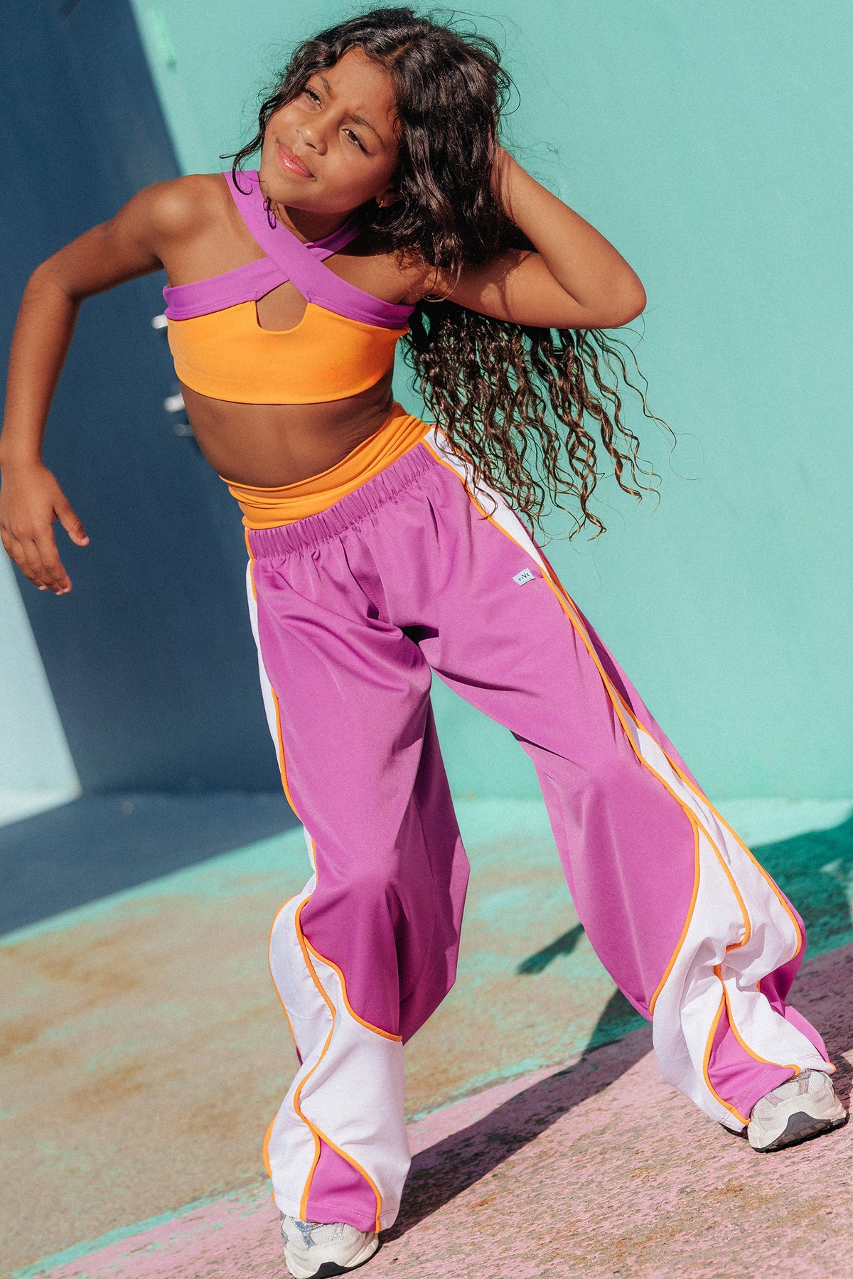 kids,  young girl posing in a vibrant purple and orange athletic crop top with matching purple sweatpants featuring white side panels and orange piping, set against a colorful teal and pink wall.