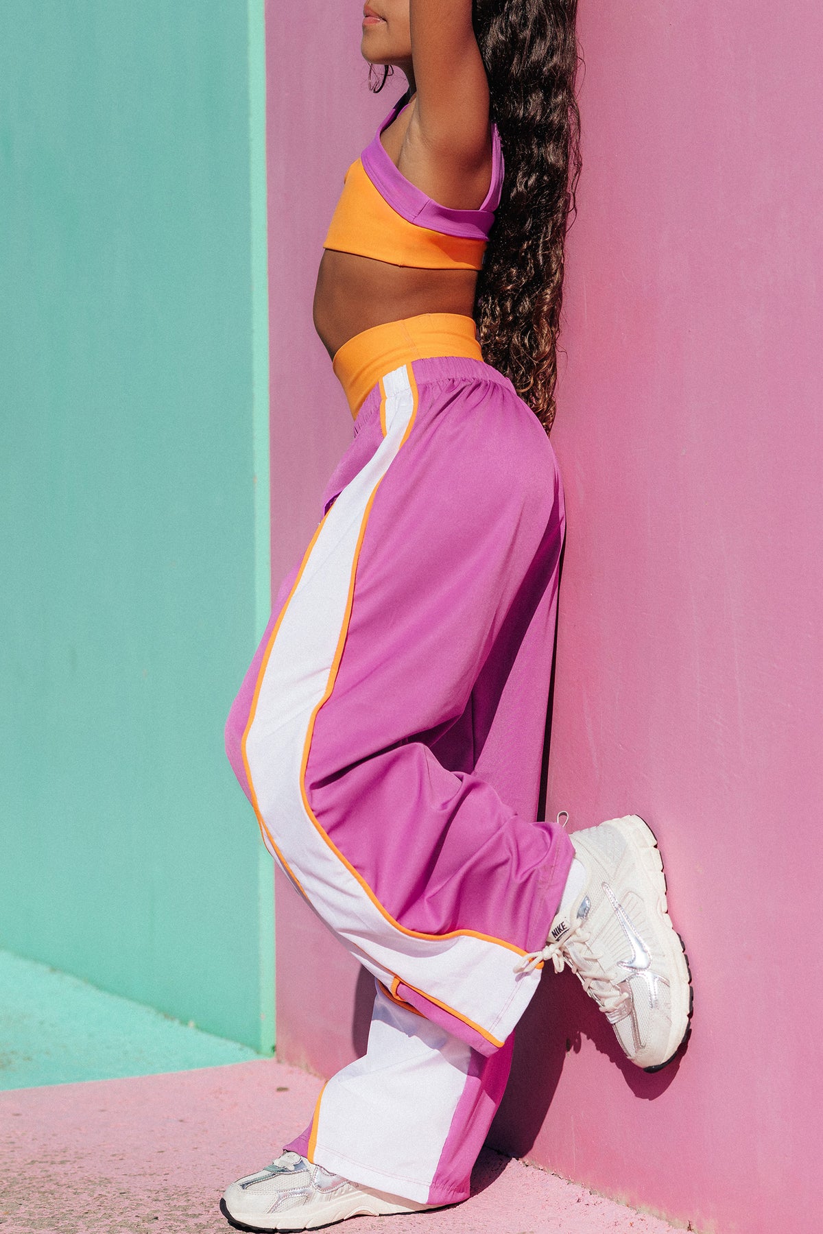 kids, A person wearing a bright pink and orange athletic crop top with matching pink and orange wide-leg track pants, paired with white sneakers, leaning against a pink wall.