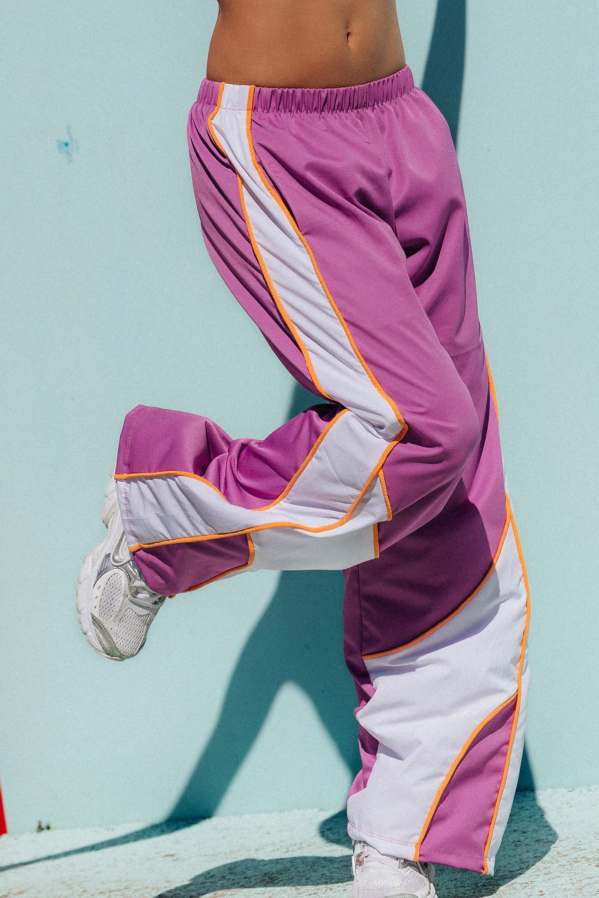 teen, A person wearing loose purple athletic track pants with large white side panels and thin orange piping, photographed outdoors against a light blue wall.