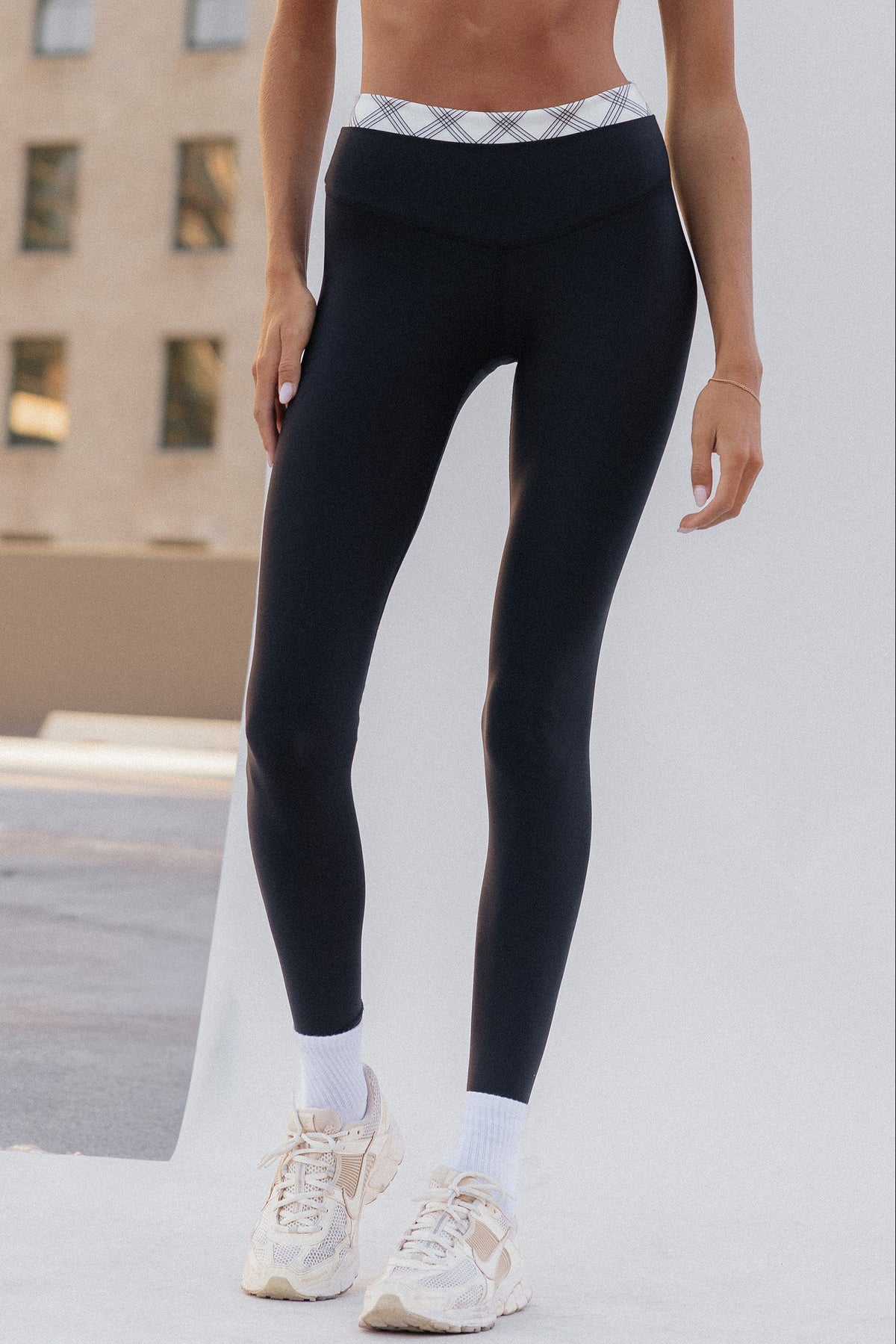 teen, wearing black high-waisted leggings with a white and gray plaid patterned waistband, paired with white socks and beige sneakers, standing outdoors against a light-colored wall.