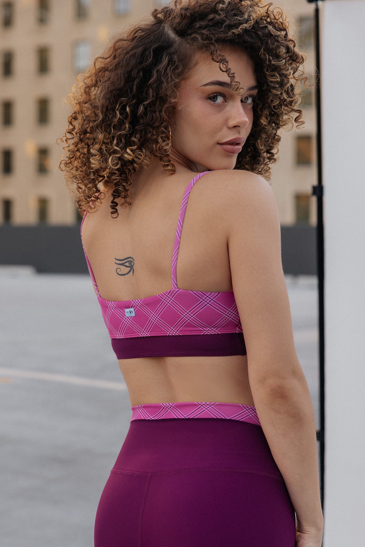 women, A woman with curly brown hair is shown from the back wearing a pink and purple athletic sports bra and matching high-waisted leggings. The setting appears to be an outdoor rooftop or urban location with a building in the background.