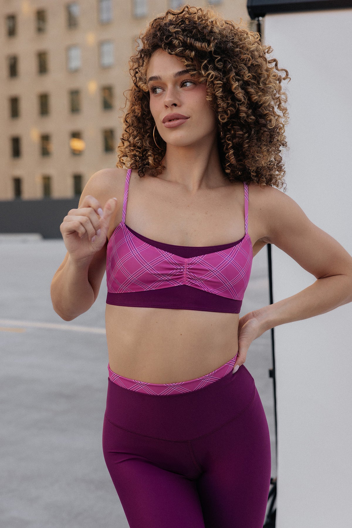 women, A woman with curly brown hair wearing a matching pink and purple athletic set, featuring a ruched sports bra and high-waisted leggings, posing outdoors against a white studio backdrop with a building in the background.