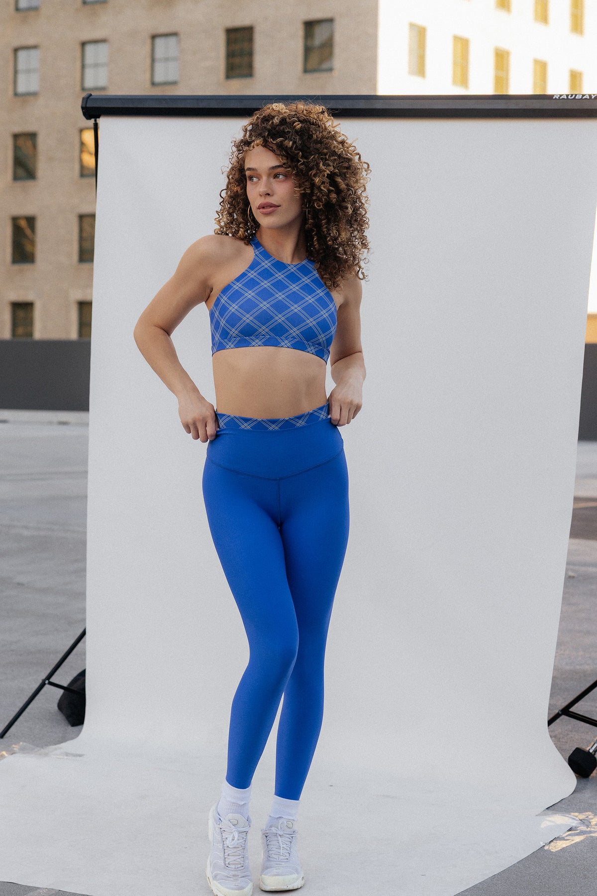 women, A woman modeling a matching athletic set consisting of a blue patterned sports bra and bright blue leggings, standing in front of a white studio backdrop with buildings visible in the background.

