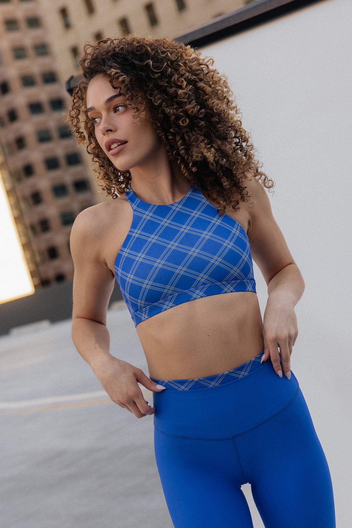women, A woman with curly brown hair wearing a blue plaid sports bra and matching blue leggings, posing outdoors against a white wall with a building in the background.