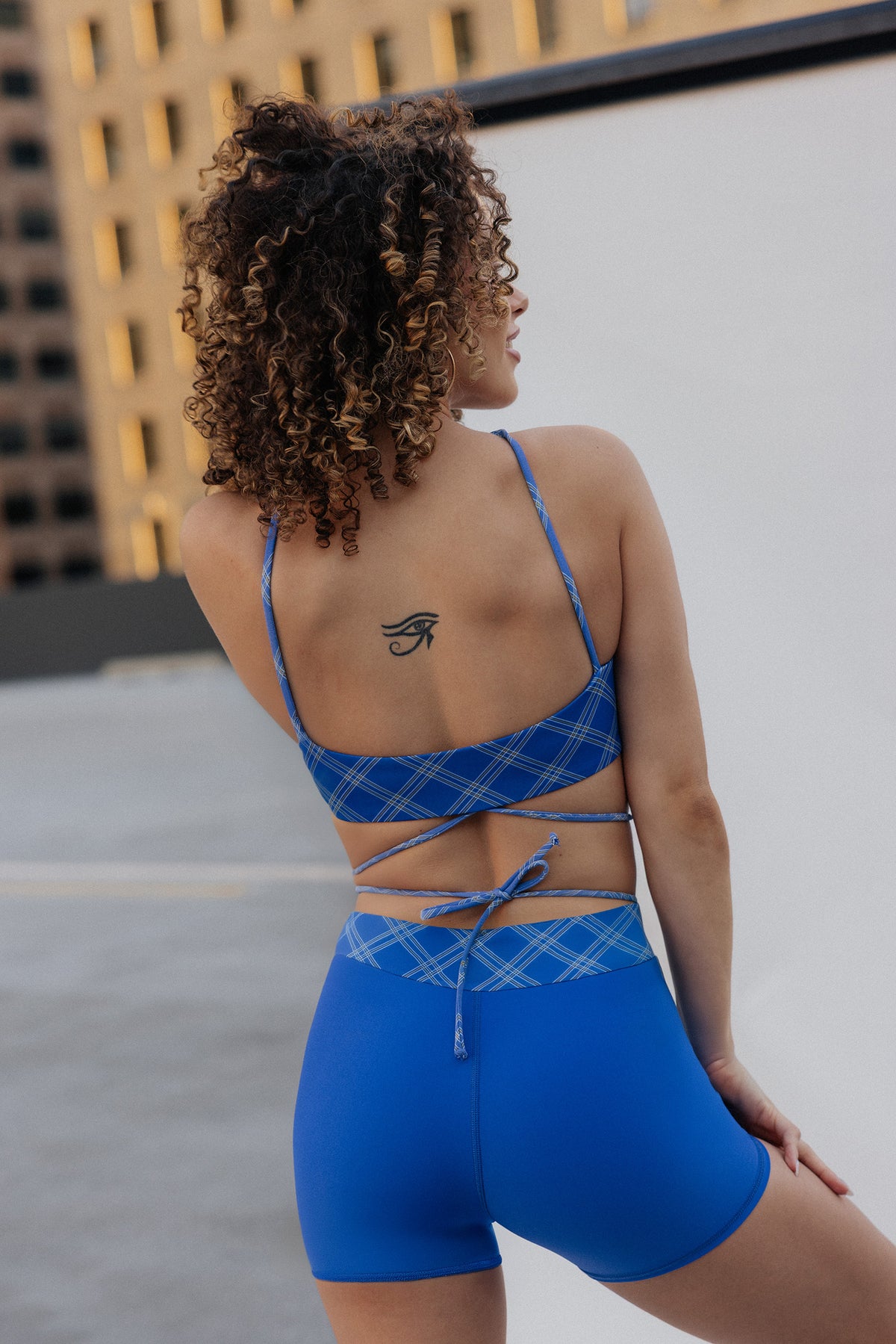 women, A woman with curly hair is shown from behind wearing a blue plaid athletic set consisting of a strappy crop top and matching high-waisted shorts, set against a modern outdoor background.