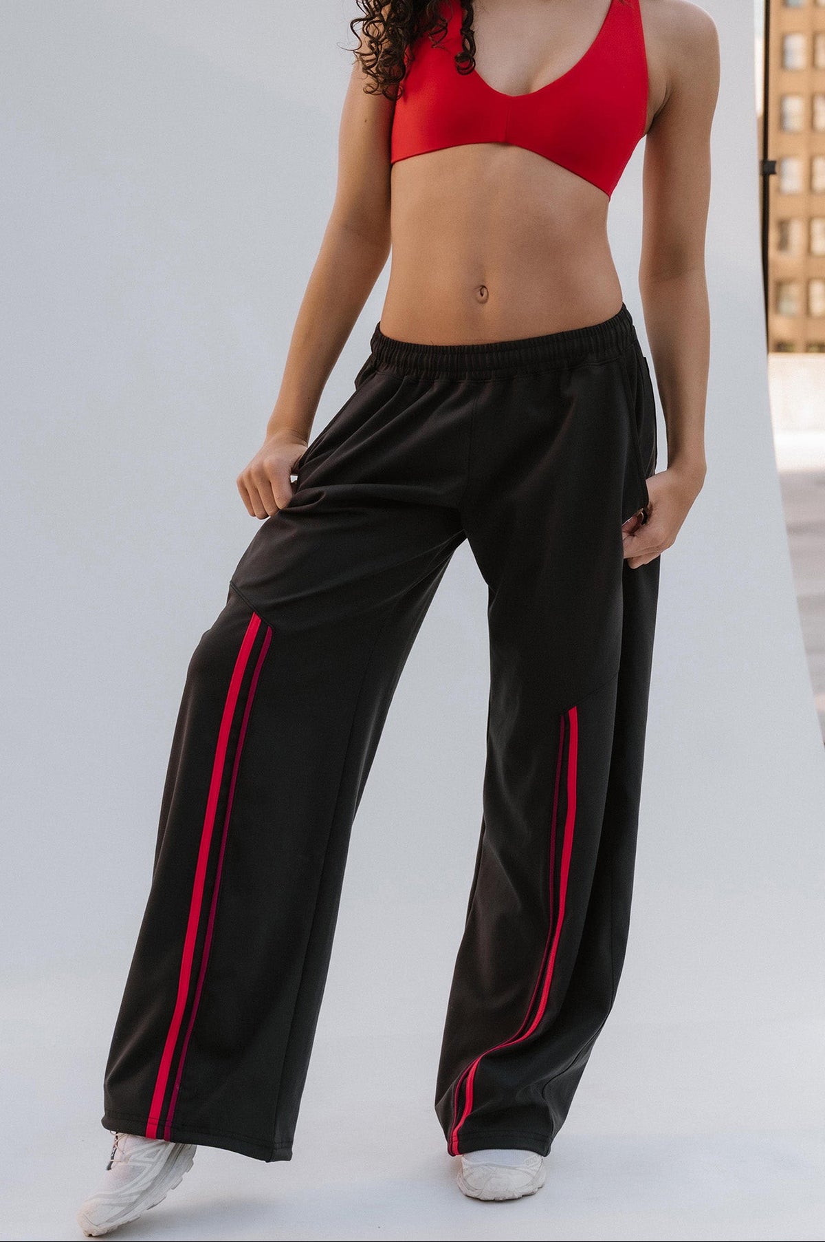 teen, modeling a red sports bra and black wide-leg sweatpants with red side stripes, standing on a white studio sweep with city buildings in the background.