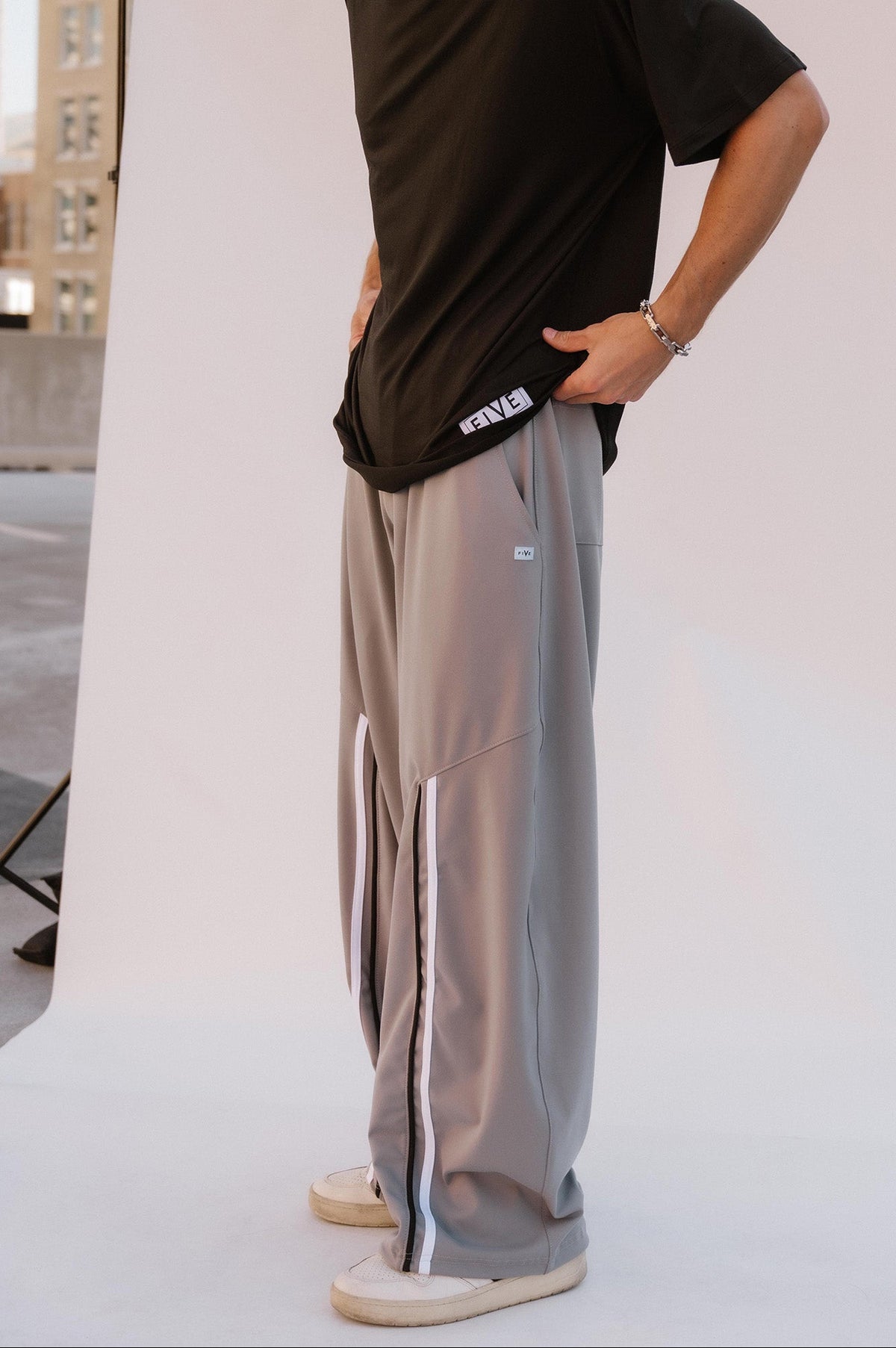 men, A man wearing a black t-shirt and light gray wide-leg sweatpants with a white stripe, standing in front of a white studio backdrop with city buildings in the background.