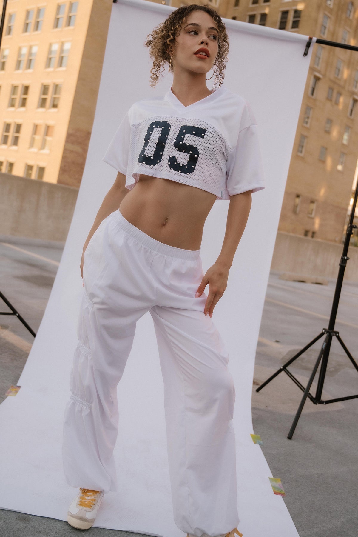 A model wearing a white cropped t-shirt with '05' in black sequins and matching white sweatpants, standing in front of a white studio backdrop on a rooftop with buildings in the background.