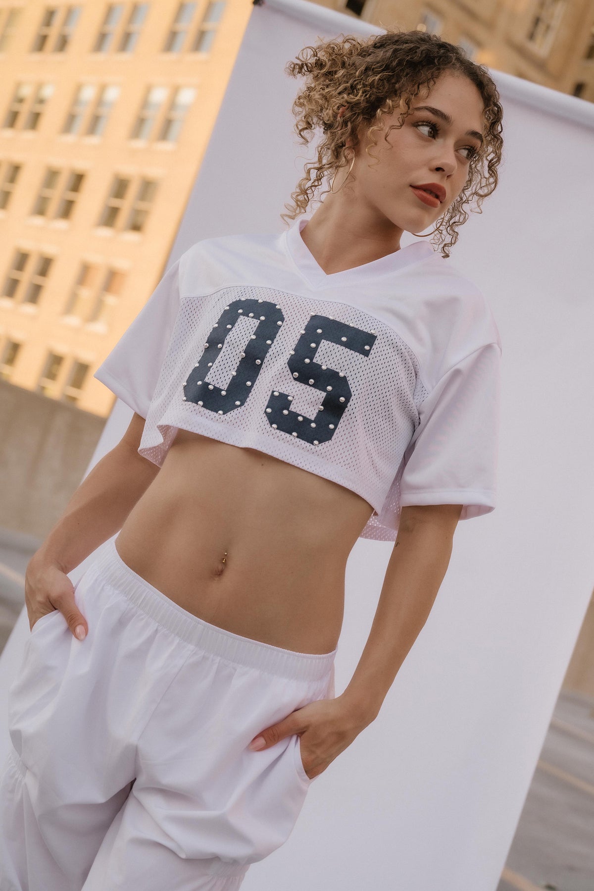 A woman wearing a white cropped mesh football-style jersey with large dark navy '05' lettering on the front, paired with white sweatpants, posed in front of a white studio backdrop with a city building in the background.