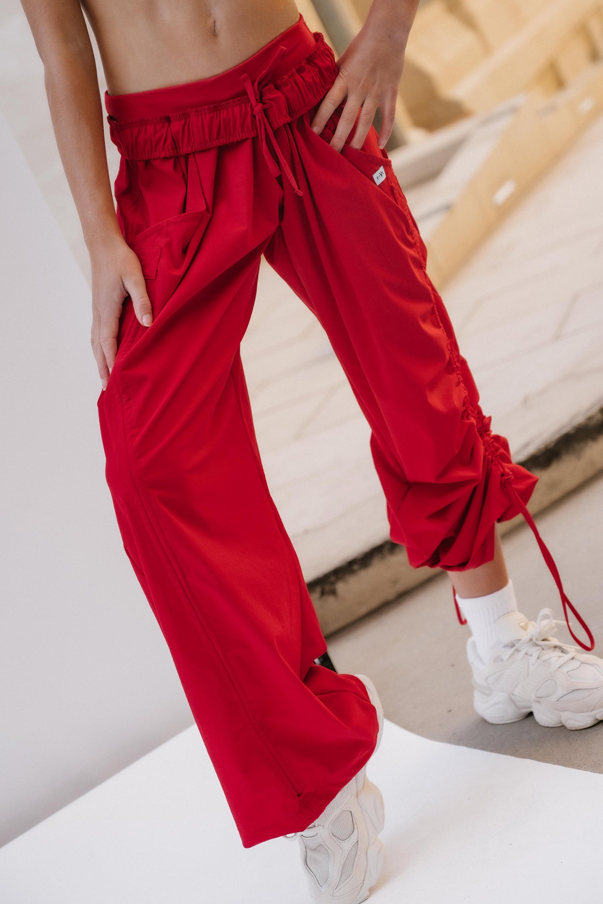 kids, A person wearing bright red wide-leg drawstring pants with gathered cuffs and white sneakers, photographed in a minimalist indoor setting.