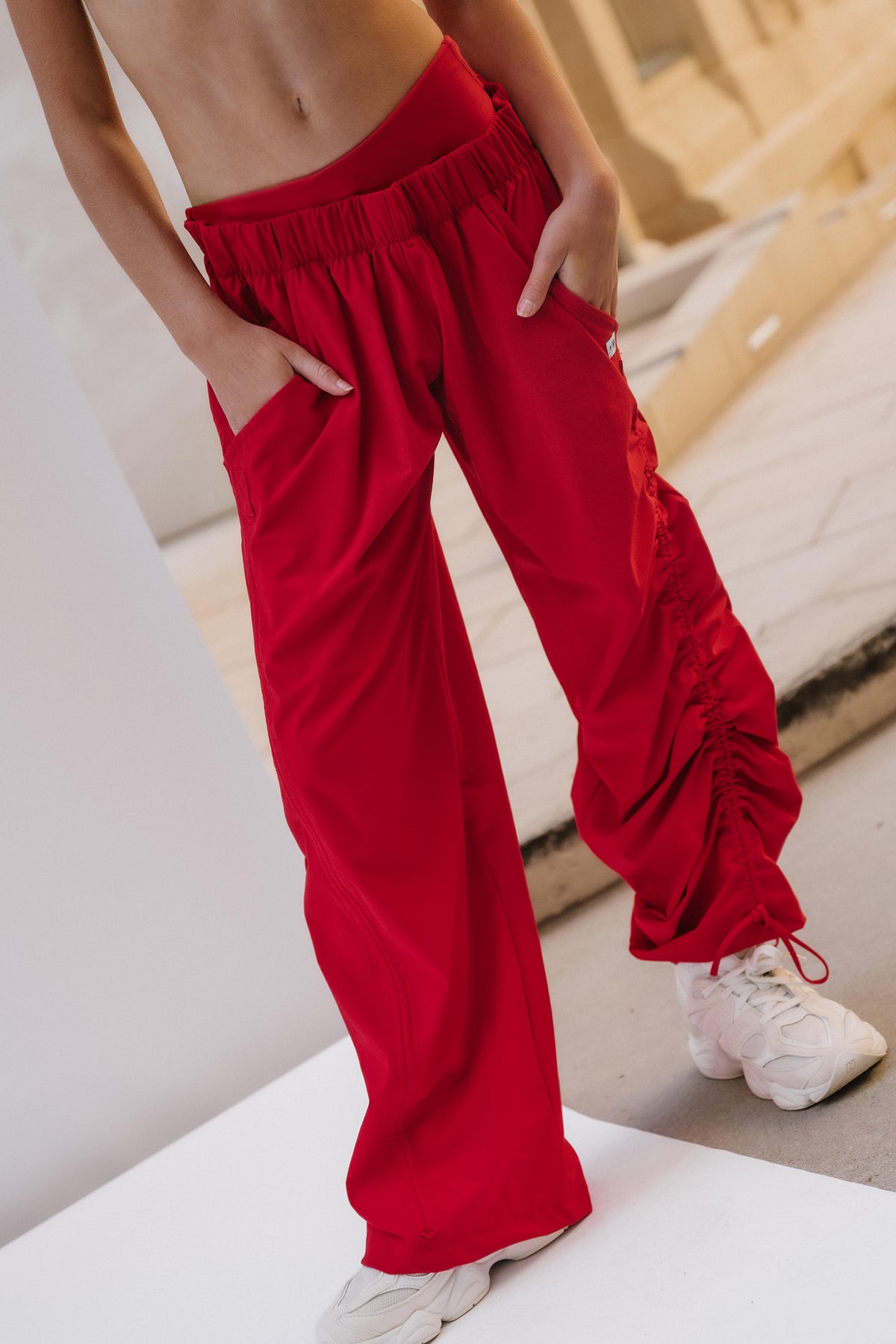 kids, A person wearing bright red wide-leg drawstring pants with side pockets and gathered cuffs, paired with white sneakers, standing in a minimalist indoor setting.