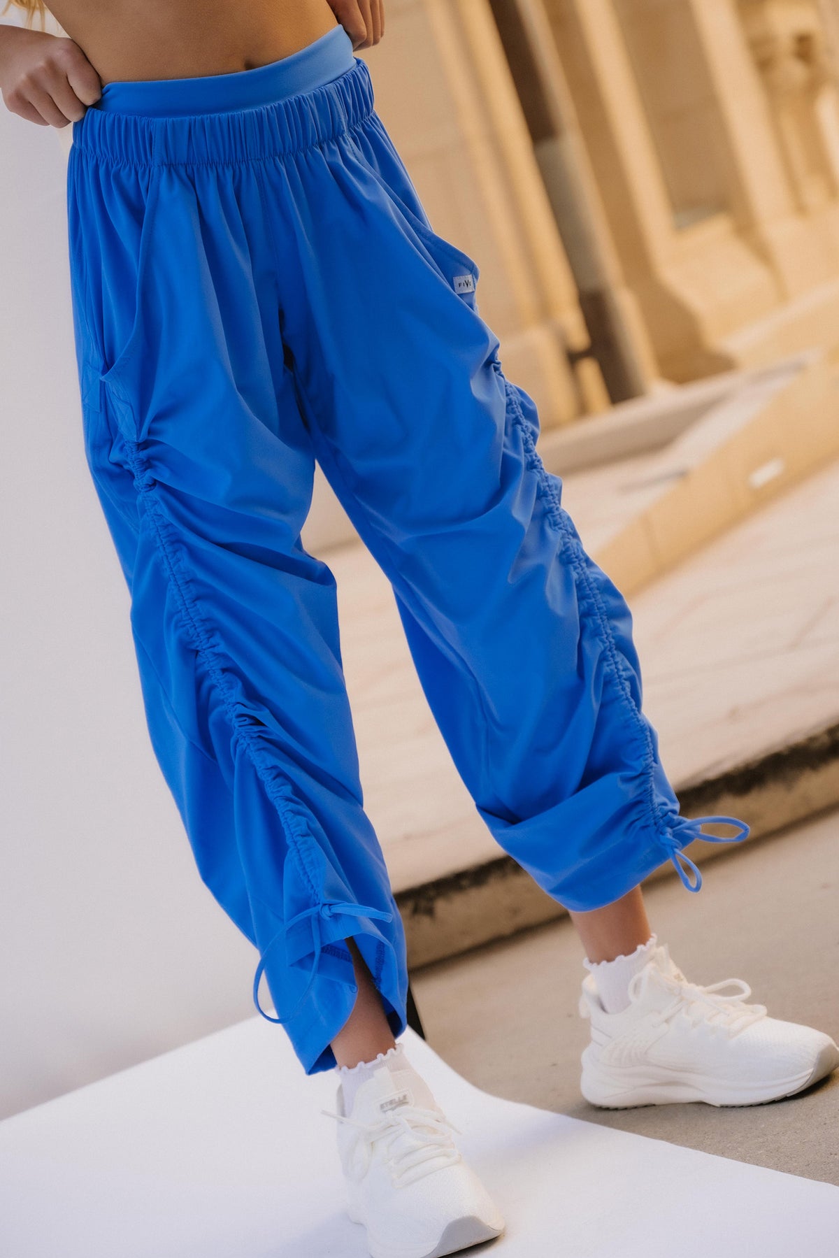 kids, A model wearing bright blue ruched ruched sweatpants, styled with a matching blue sports bra and white sneakers, standing on a white platform.