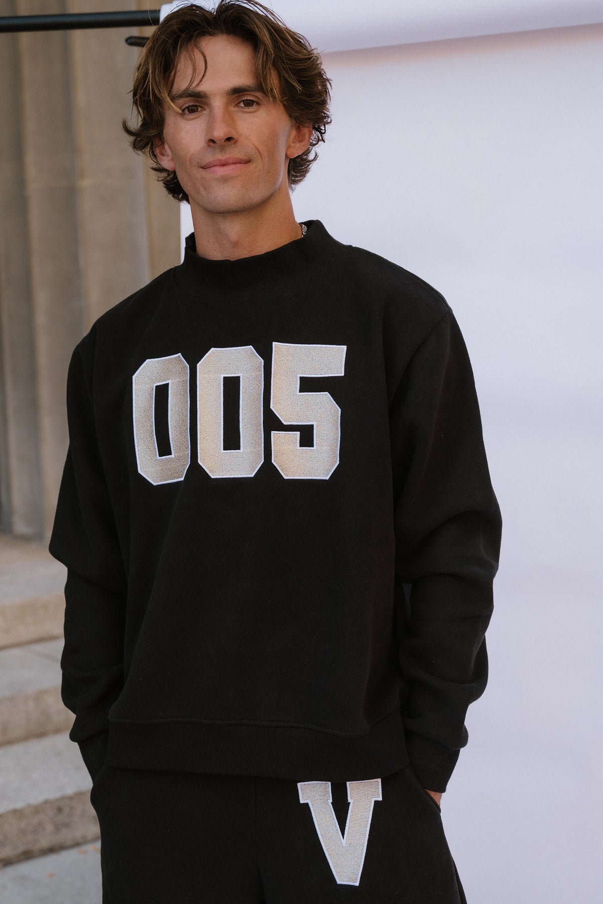 mens, A man wearing a black crewneck sweatshirt with large white block numbers '005' on the front, paired with black sweatpants featuring a white 'V' on the thigh, set against a light gray background.